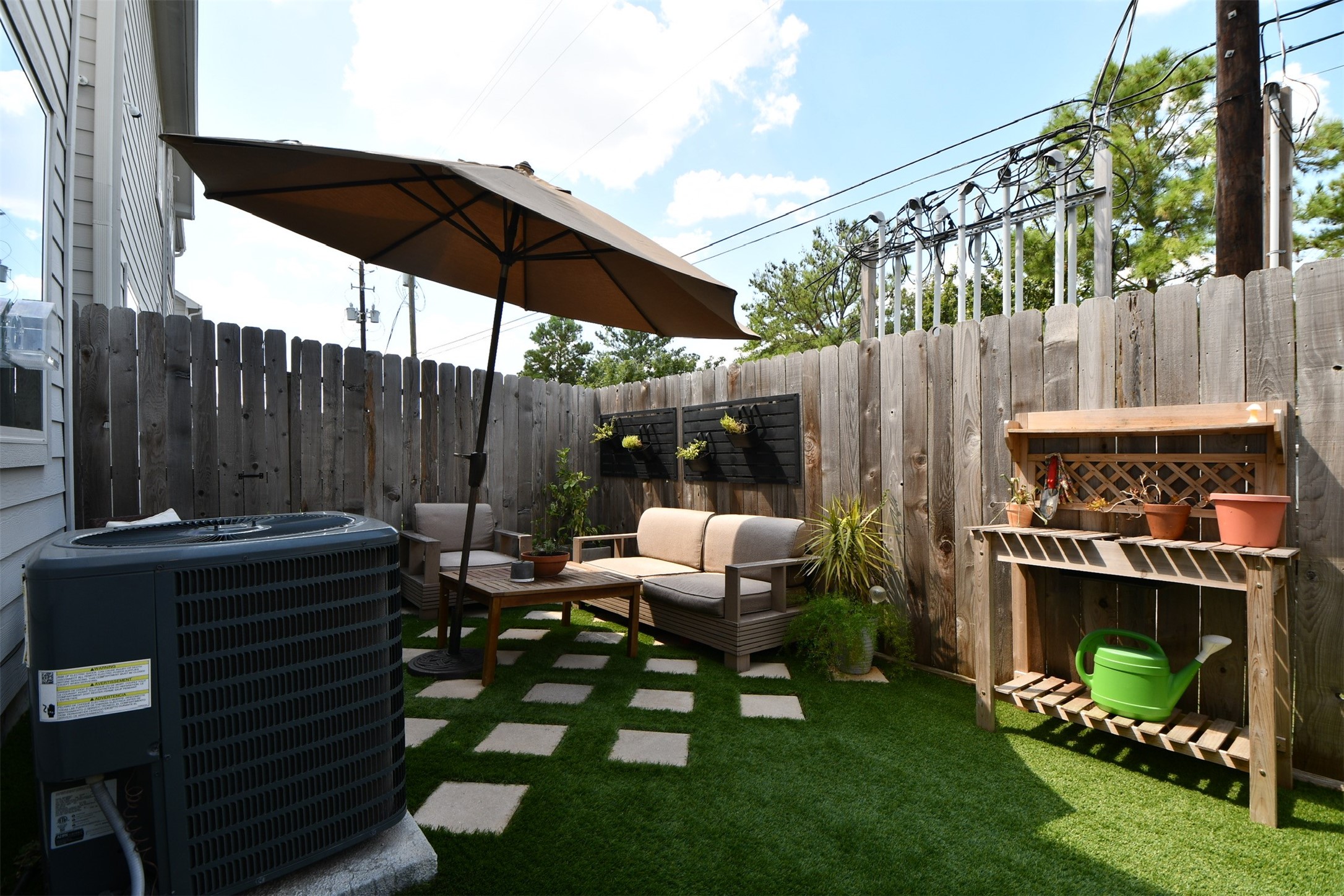 1239 West Tidwell Road Houston, TX 77091 - Photo 34 of 35 Cozy backyard patio with artificial grass, featuring a small seating area under a large umbrella, surrounded by a wooden fence. There's a gardening station and potted plants, creating a relaxing outdoor space.