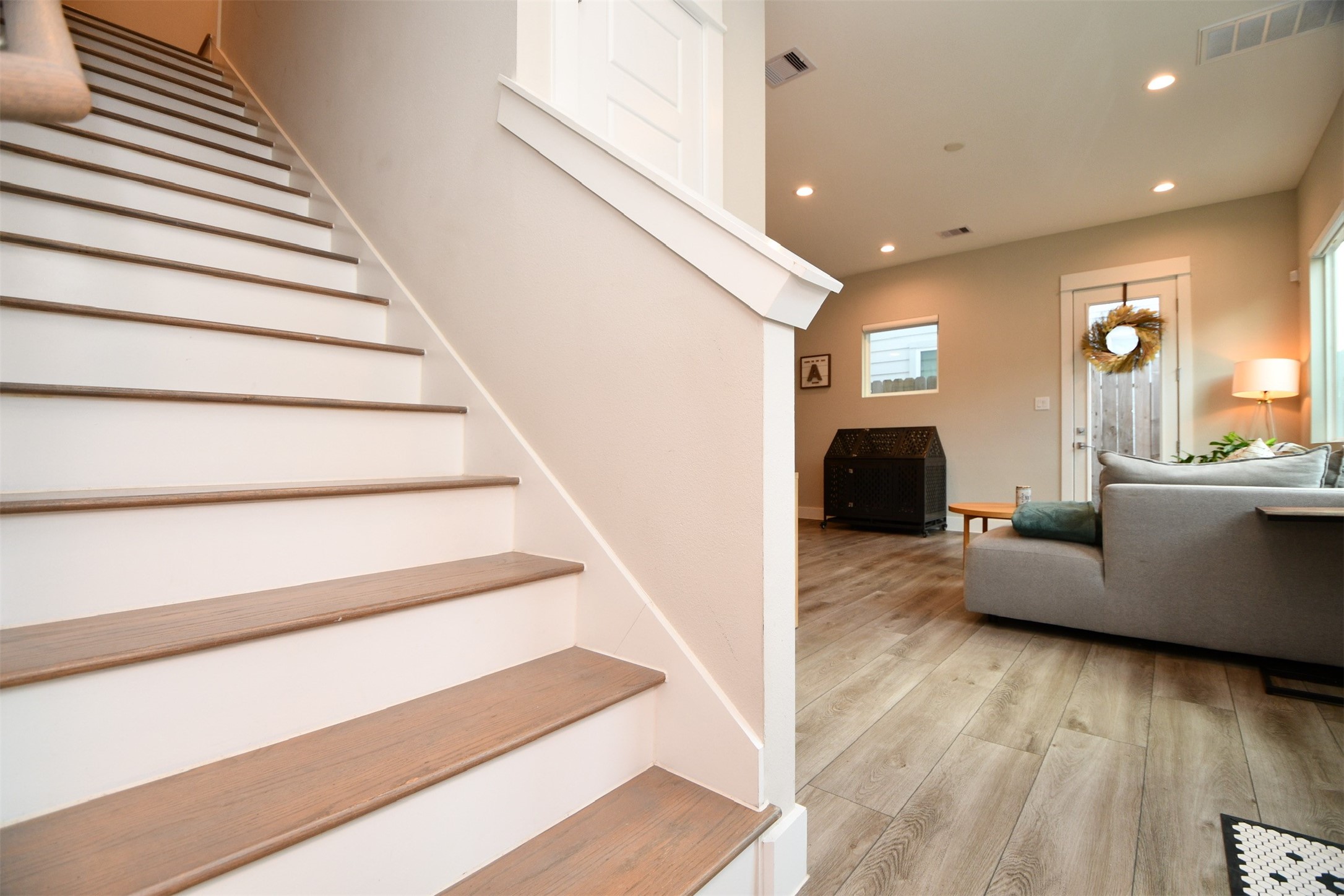 1239 West Tidwell Road Houston, TX 77091 - Photo 6 of 35 This photo showcases a modern living area with wood-style flooring, a cozy gray sofa, and a well-lit ambiance. There's a welcoming staircase with wooden steps and a glimpse of a door adorned with a wreath, adding a touch of charm.