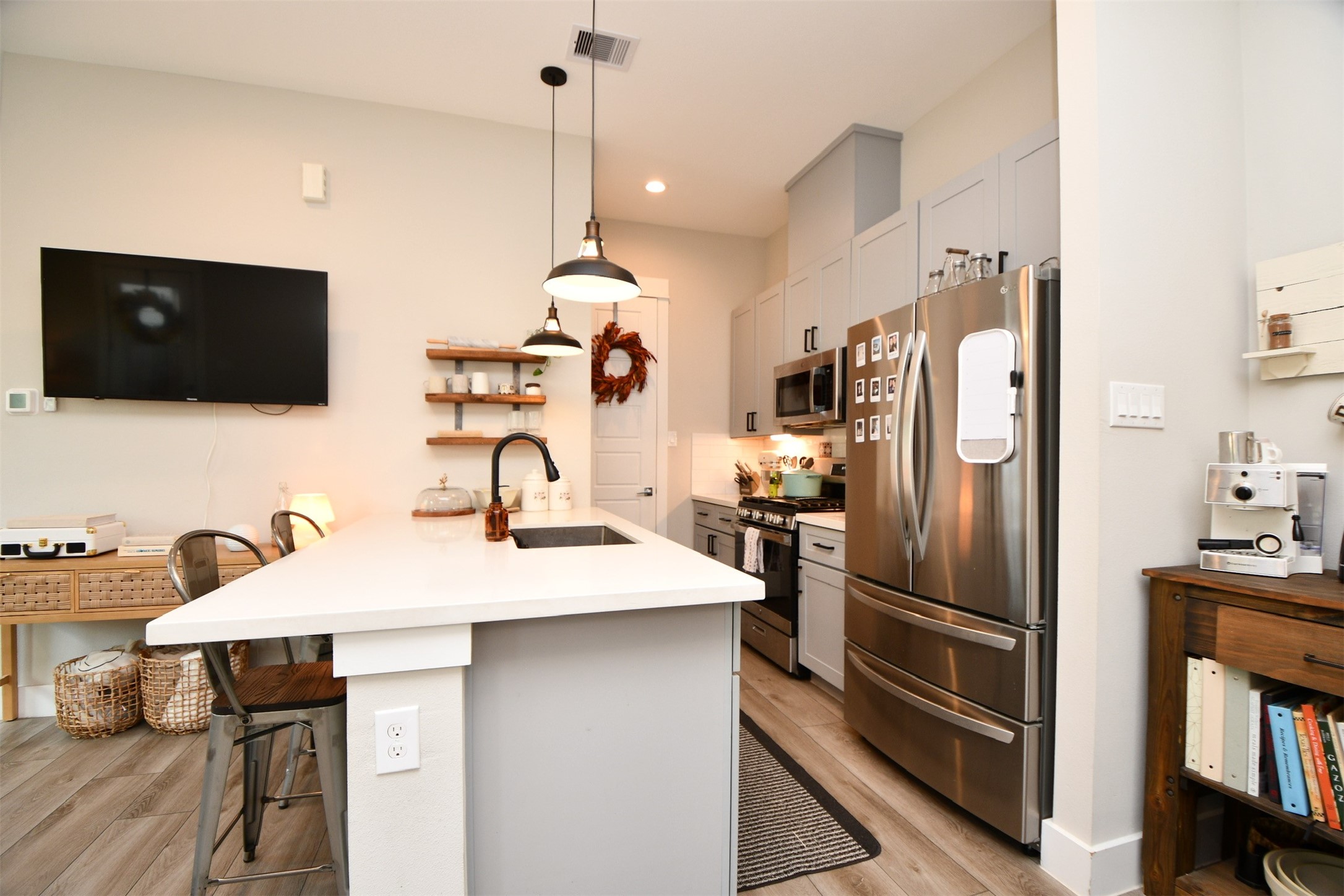 1239 West Tidwell Road Houston, TX 77091 - Photo 9 of 35 This photo showcases a modern kitchen with stainless steel appliances, white cabinetry, and a central island with seating. The space is well-lit with pendant lights, featuring a cozy and functional design. A mounted TV and a small dining area add to the room's versatility.