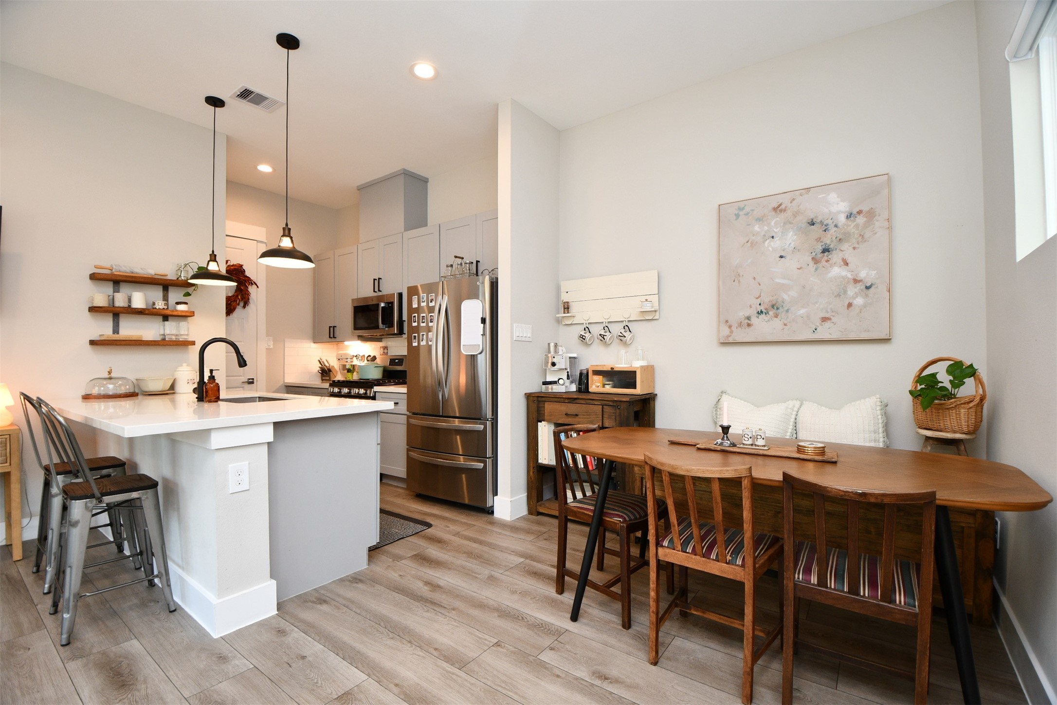 1239 West Tidwell Road Houston, TX 77091 - Photo 10 of 35 This photo showcases a modern kitchen and dining area with light wood flooring. The kitchen features a sleek design with a center island, pendant lighting, stainless steel appliances, and open shelving. The adjacent dining area has a cozy wooden table with chairs, accented by wall art and a small plant, creating a warm and inviting space.