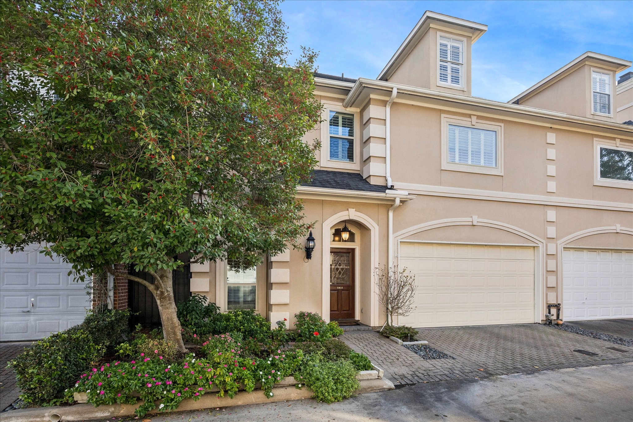 This charming townhouse features a classic beige exterior with white accents and a welcoming front door. A mature tree and lush landscaping enhance the curb appeal, while a spacious driveway leads to an attached garage.