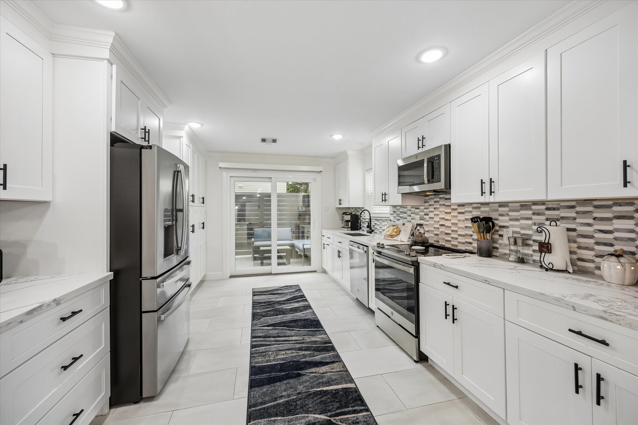 1420 Indiana Street Houston, TX 77006 - Photo 11 of 31 This modern kitchen features sleek white cabinetry, stainless steel appliances, and a stylish tile backsplash. It has ample counter space, and sliding glass doors leading to an outdoor area, creating a bright and inviting atmosphere.