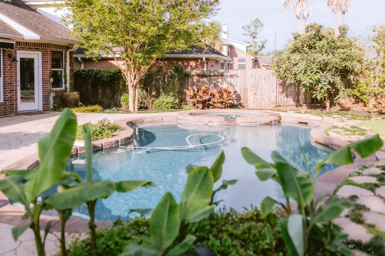 22402 Rue Canyon Court Katy, TX 77450 - Photo 16 of 18 Framed beautifully by broad banana leaves, this shot showcases the vibrant, healthy landscaping that sets this backyard apart. The deep blue pool and integrated hot tub are perfectly complemented by shaded retreat areas under mature trees, making it an idyllic sanctuary for both playful pool days and quiet afternoons with a book.