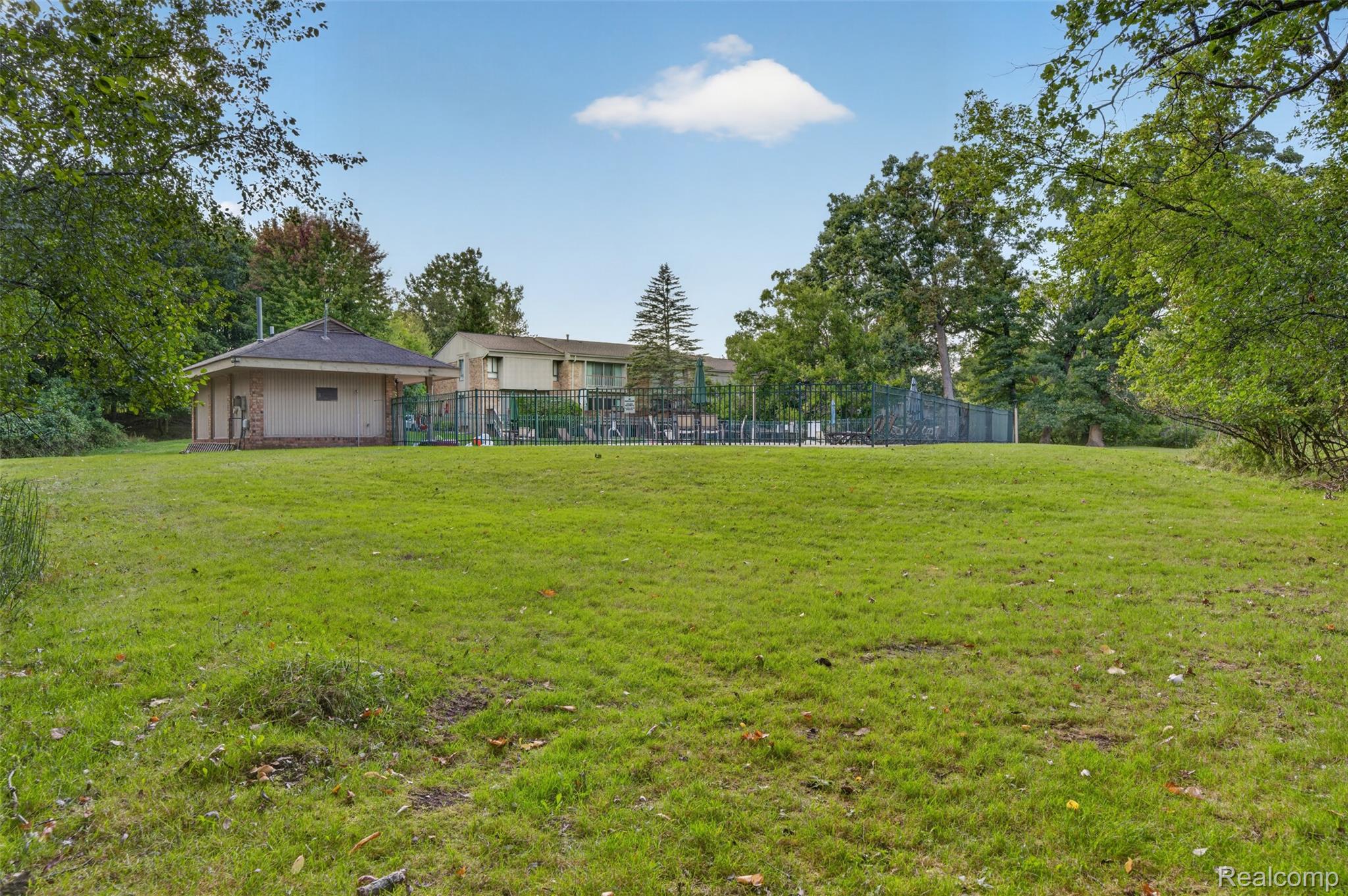 1439 Oakbrook E Drive Rochester, MI 48307 - Photo 8 of 68 Expansive grassy area with mature perimeter trees, a nearby brick-clad utility building with an asphalt shingle roof, and a visible swimming pool area with a metal fence