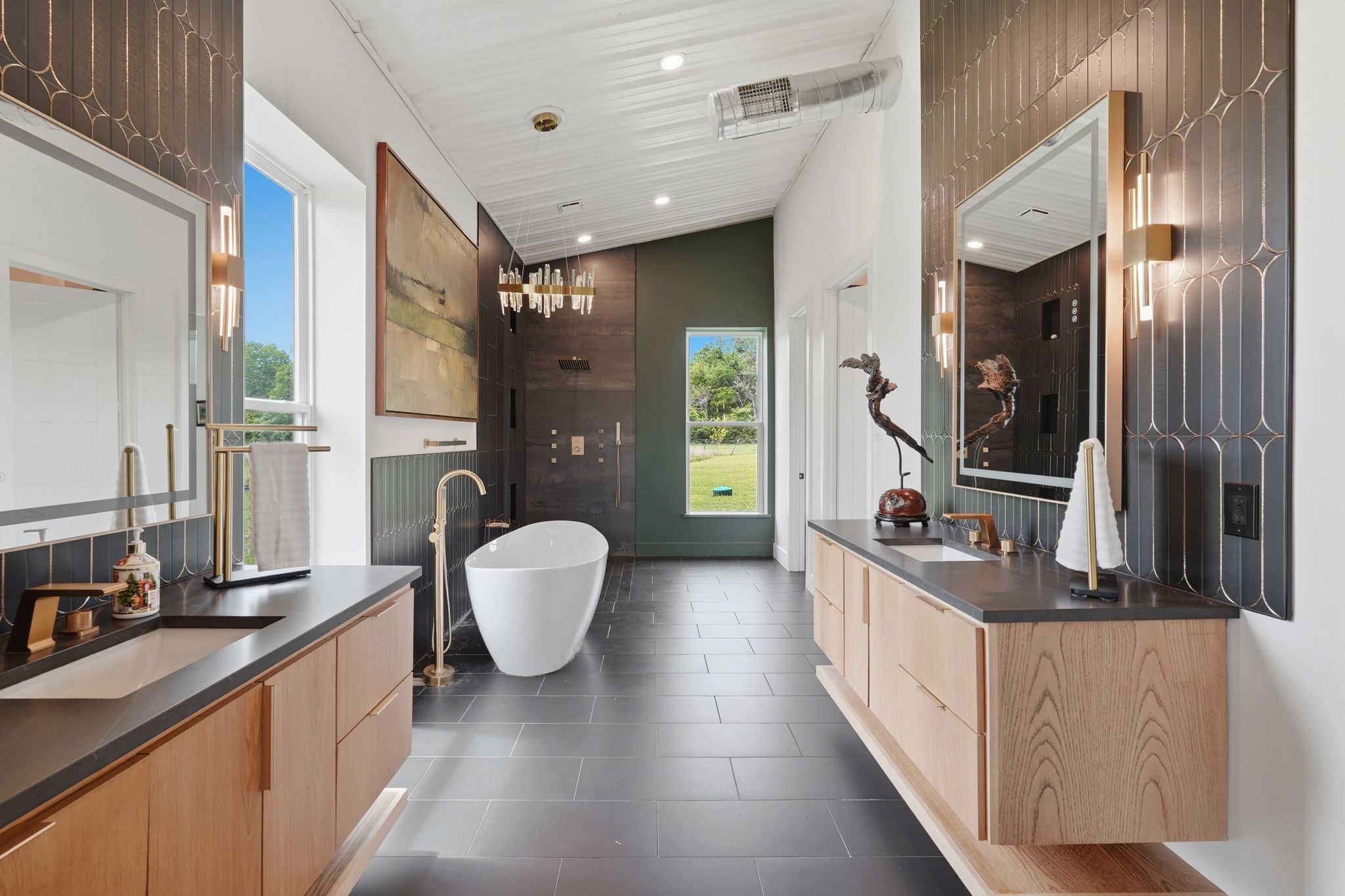 2743 Bobville Road Montgomery, TX 77316 - Photo 29 of 50 This bathroom features a modern design with sleek black tile flooring, dual wooden vanities with black countertops, and contemporary lighting. A freestanding bathtub sits centrally, with a large window offering natural light and views of greenery. The shower area is accented by dark tiles and a stylish chandelier.