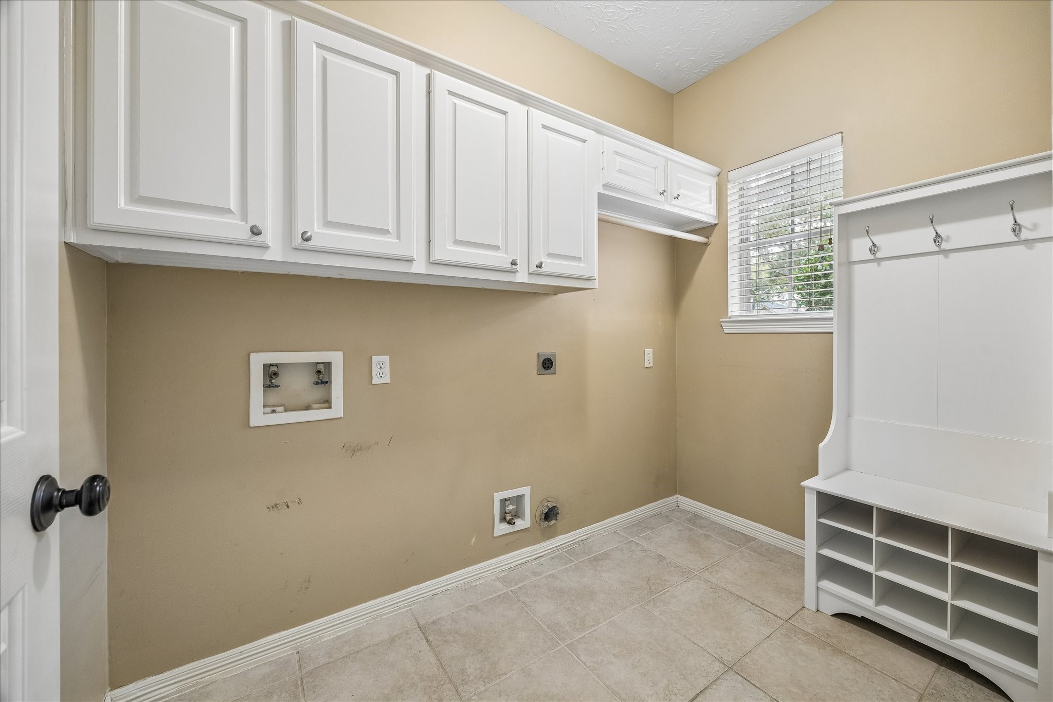 5310 Summerland Ridge Court Houston, TX 77041 - Photo 38 of 46 This is a bright laundry room with beige walls and white cabinetry. It features hookups for a washer and dryer, a window for natural light, and a white storage bench with hooks and cubbies.