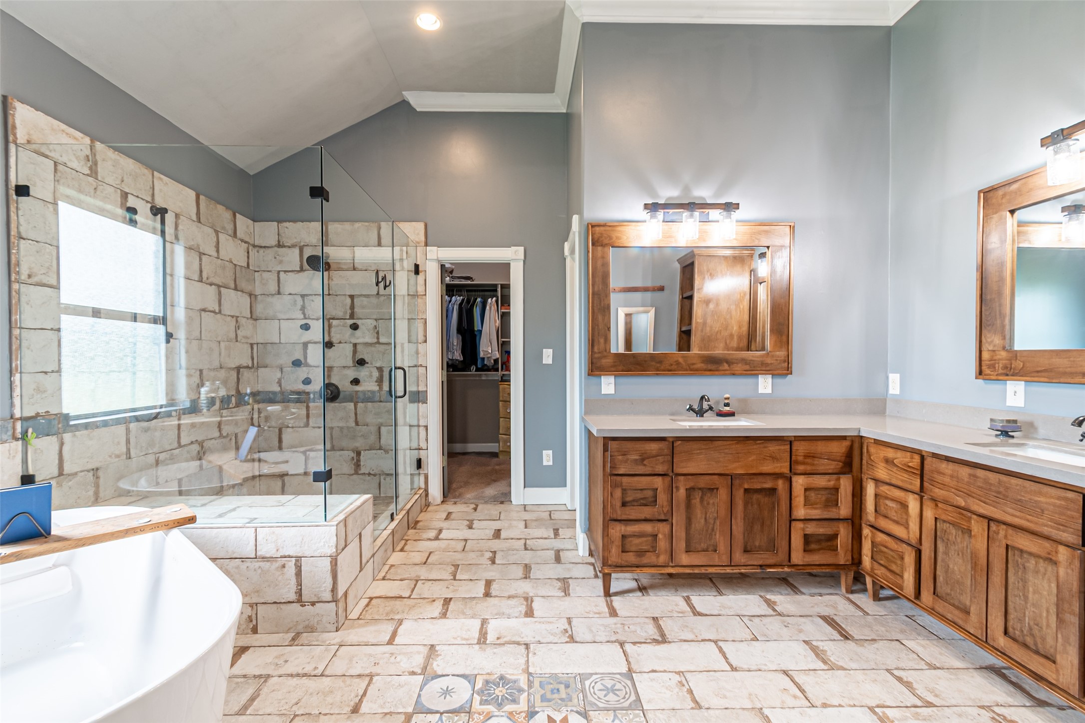 38766 Wyatt Chapel Road Hempstead, TX 77445 - Photo 9 of 45 This spacious bathroom features a large glass-enclosed shower with rustic stone tiles, dual wooden vanities with modern lighting, and a separate bathtub. It offers a cozy, elegant feel with plenty of natural light and direct access to a walk-in closet.