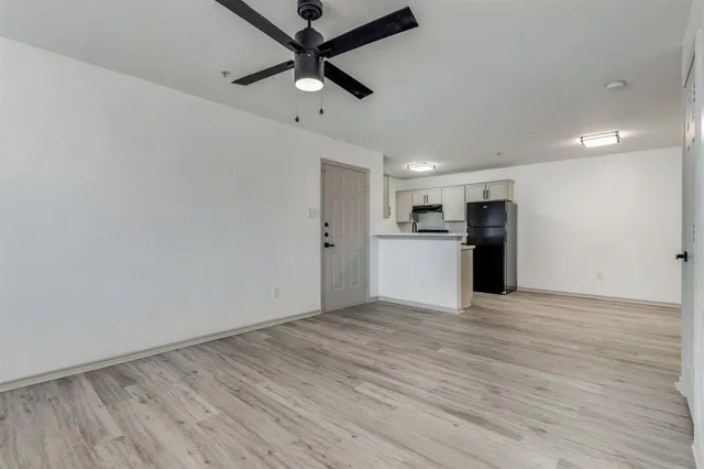 an empty room with wooden floor a ceiling fan a kitchen view and a refrigerator