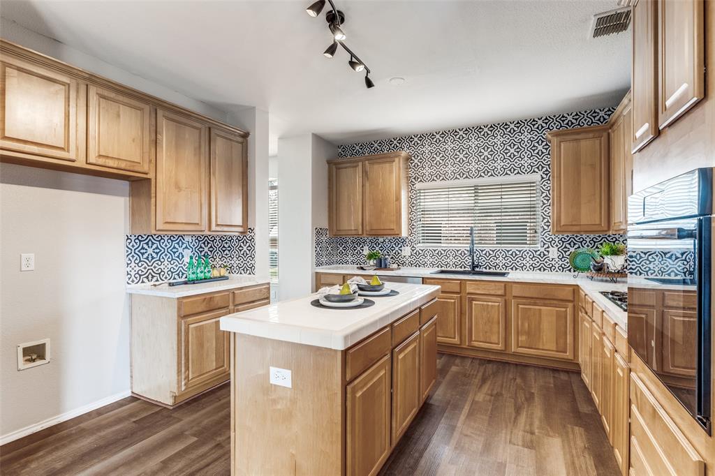 1445 Berkley Road Allen, TX 75002 - Photo 8 of 30 The island kitchen features an abundance of wood cabinetry, and was updated in 2024-25 with ceramic tile backsplash and wall accents, black sink, stainless steel dishwasher, and fresh neutral counter tops along the perimeter.