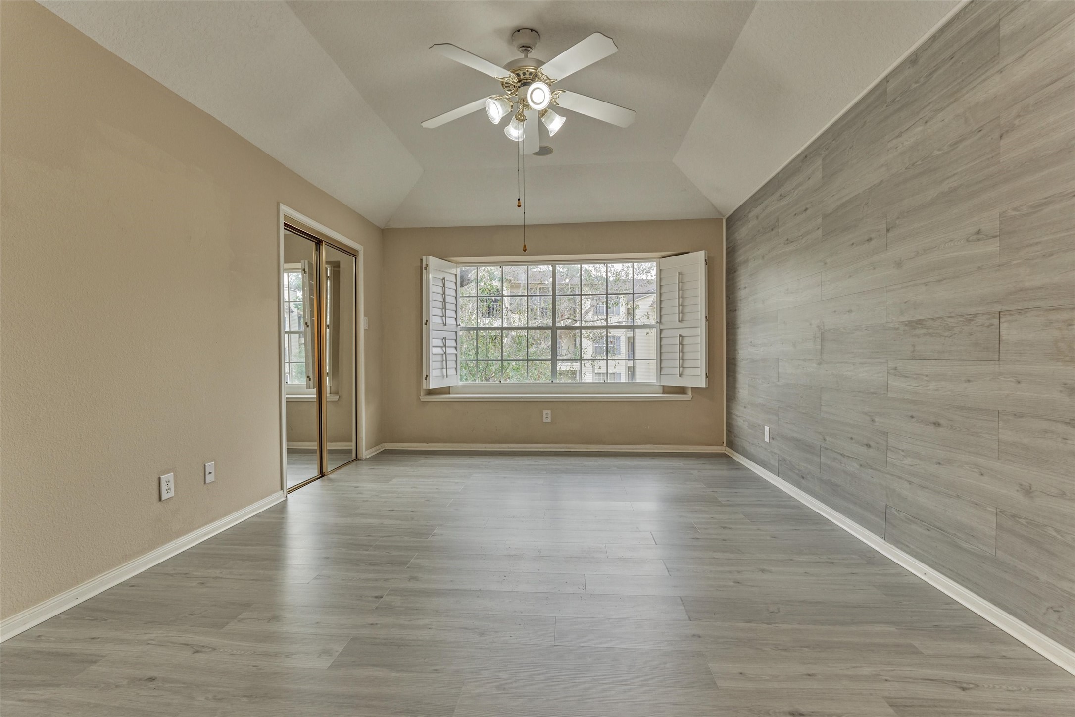 14420 Walters Road, Unit 26 Houston, TX 77014 - Photo 27 of 45 The primary bedroom features a vaulted ceiling with a ceiling fan, large windows with shutters for natural light, light wood-look flooring, and a mirrored closet. The neutral tones provide a versatile space ready for personal touches.