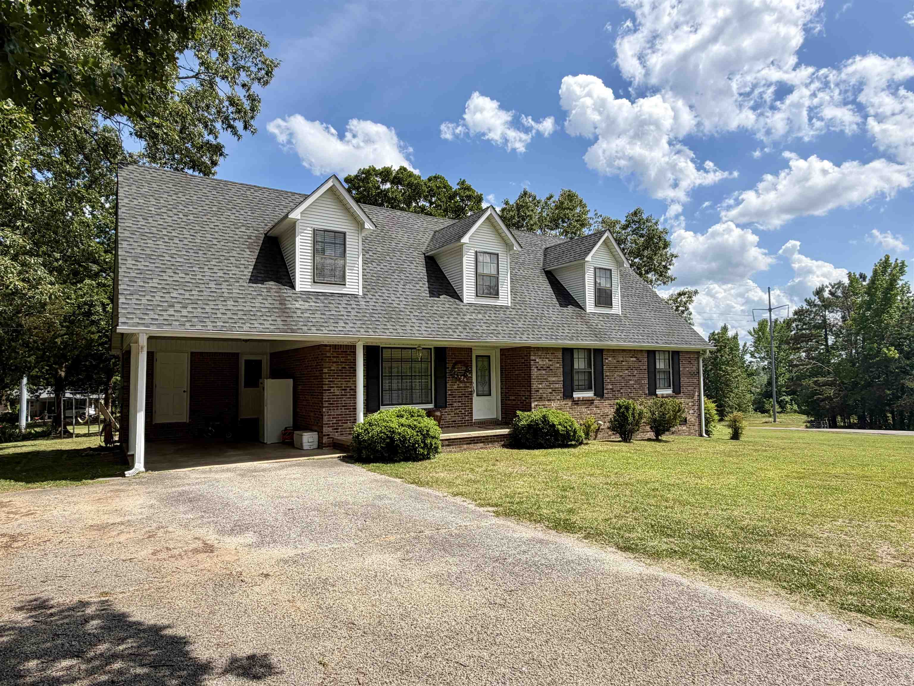 142 Treece Street Selmer, TN 38375 - Photo 1 of 36 Brick facade residence featuring a covered carport, multiple dormer windows, a gray shingle roof, and a mature tree backdrop