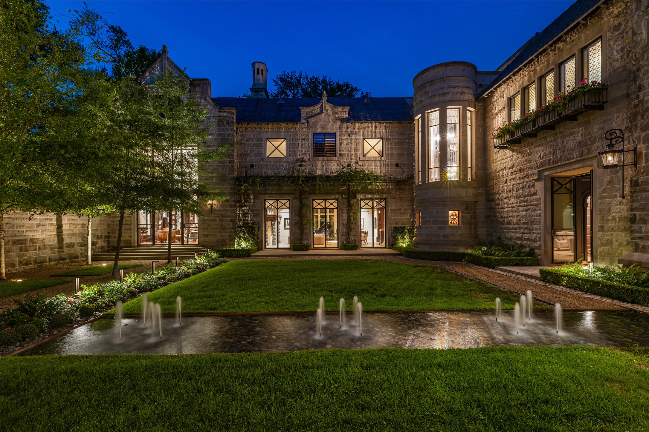 107 Timberwilde Lane Houston, TX 77024 - Photo 38 of 40 In the evening The Lodge at Hunters Creek glows at dusk, showcasing Indiana limestone, arched entry, and turreted windows. The central courtyard features manicured gardens and a reflecting pool with illuminated fountains, creating a dramatic and inviting arrival. The combination of classic Richardsonian Romanesque architecture, warm interior light, and glowing waters highlights the estate’s timeless elegance and serene atmosphere.