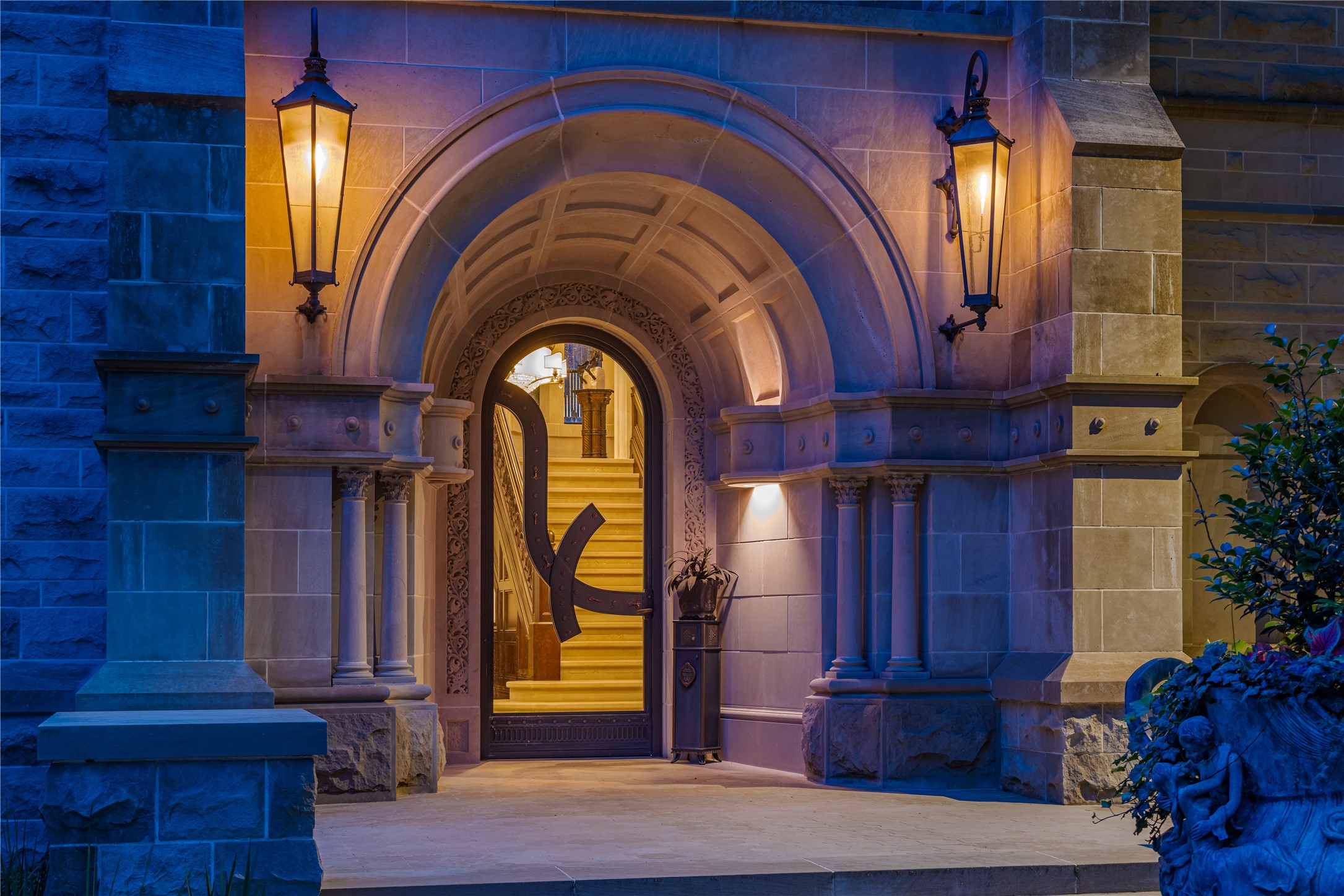 107 Timberwilde Lane Houston, TX 77024 - Photo 39 of 40 A dramatic nighttime view of the entry at The Lodge reveals the home’s monumental arched stone surround with intricate detailing, illuminated by stately gas lanterns. At its center, a striking custom bronze-and-glass contemporary door offers a bold contrast to the Richardsonian Romanesque architecture. Beyond the threshold, a softly lit staircase and refined interior reveals the grandeur within.