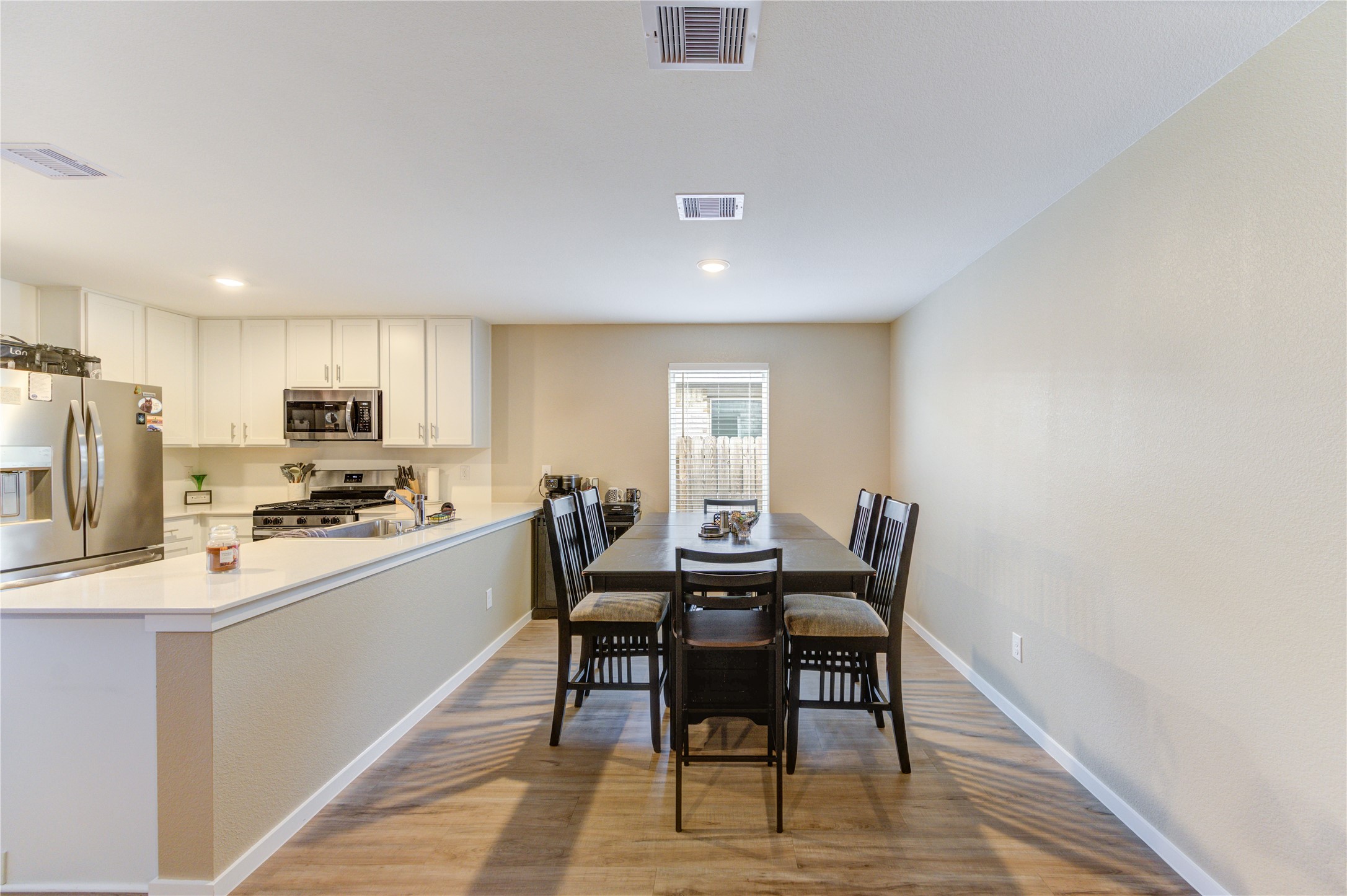 1111 Rustic Willow Drive Beasley, TX 77417 - Photo 12 of 42 This photo showcases an open-concept kitchen and dining area. The kitchen features modern stainless steel appliances, white cabinetry, and a breakfast bar. The dining area is spacious, with a large table and seating for six, highlighted by natural light from a window. The flooring is a light vinyl plank finish, enhancing the bright and airy feel of the space.