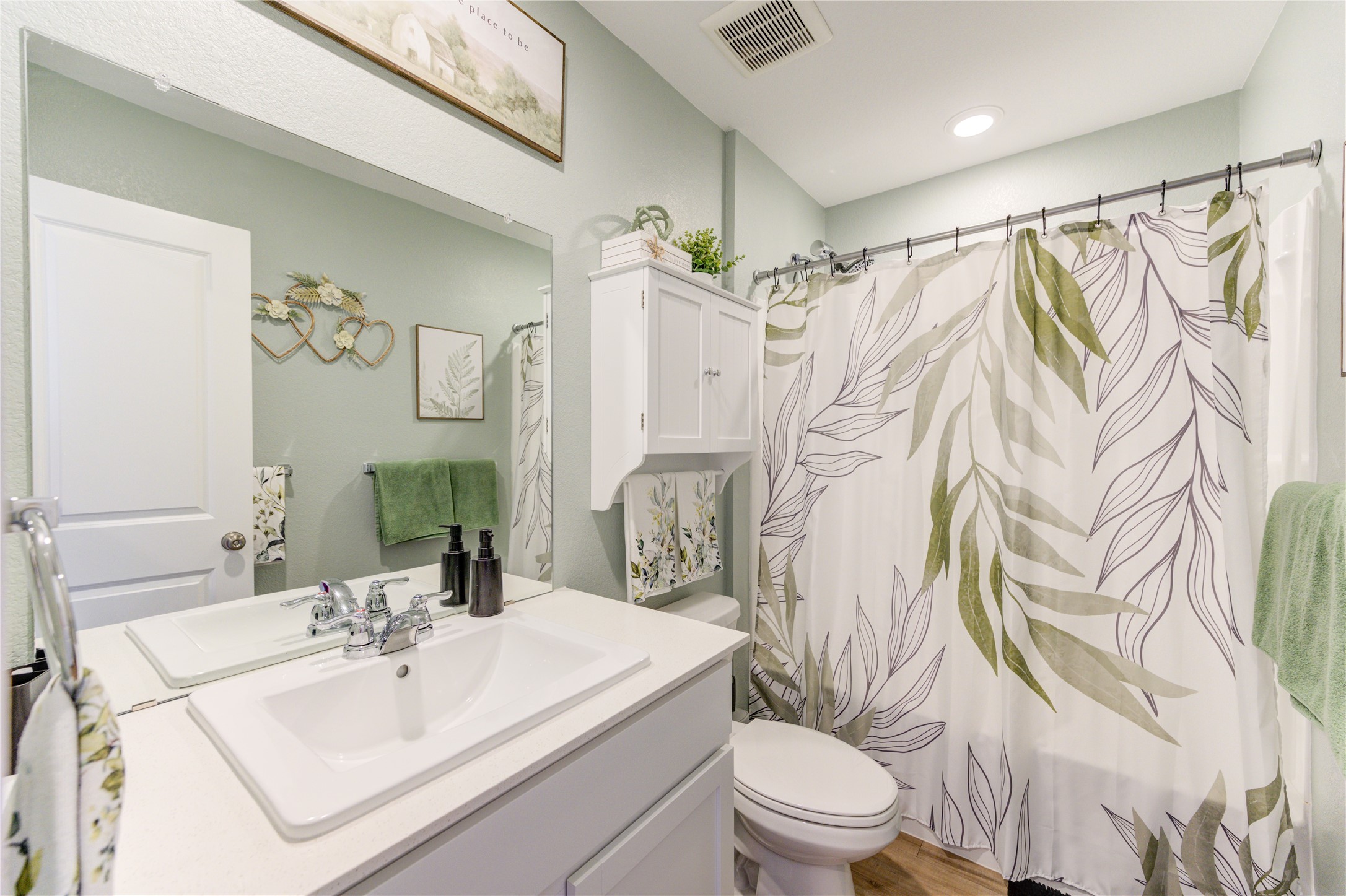 1111 Rustic Willow Drive Beasley, TX 77417 - Photo 23 of 42 This is a modern bathroom featuring a sleek white vanity with dual sinks, a stylish mirror, and ample storage. The shower has a leafy-patterned curtain, complemented by light green walls and wood look alike-style flooring, creating a fresh, inviting atmosphere.