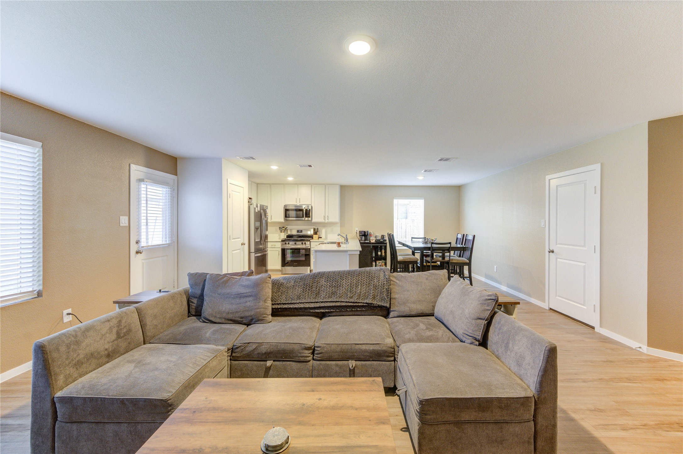 1111 Rustic Willow Drive Beasley, TX 77417 - Photo 7 of 42 This photo shows an open-concept living space with a cozy sectional sofa and a wooden coffee table. The room flows into a modern kitchen equipped with stainless steel appliances and an adjacent dining area. The space is well-lit with recessed lighting and natural light from windows, creating a warm and inviting atmosphere.