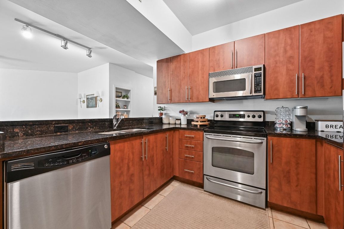 54 Rainey Street, Unit 416 Austin, TX 78701 - Photo 4 of 40 Kitchen featuring appliances with stainless steel finishes, dark stone countertops, light tile patterned flooring, and brown cabinetry