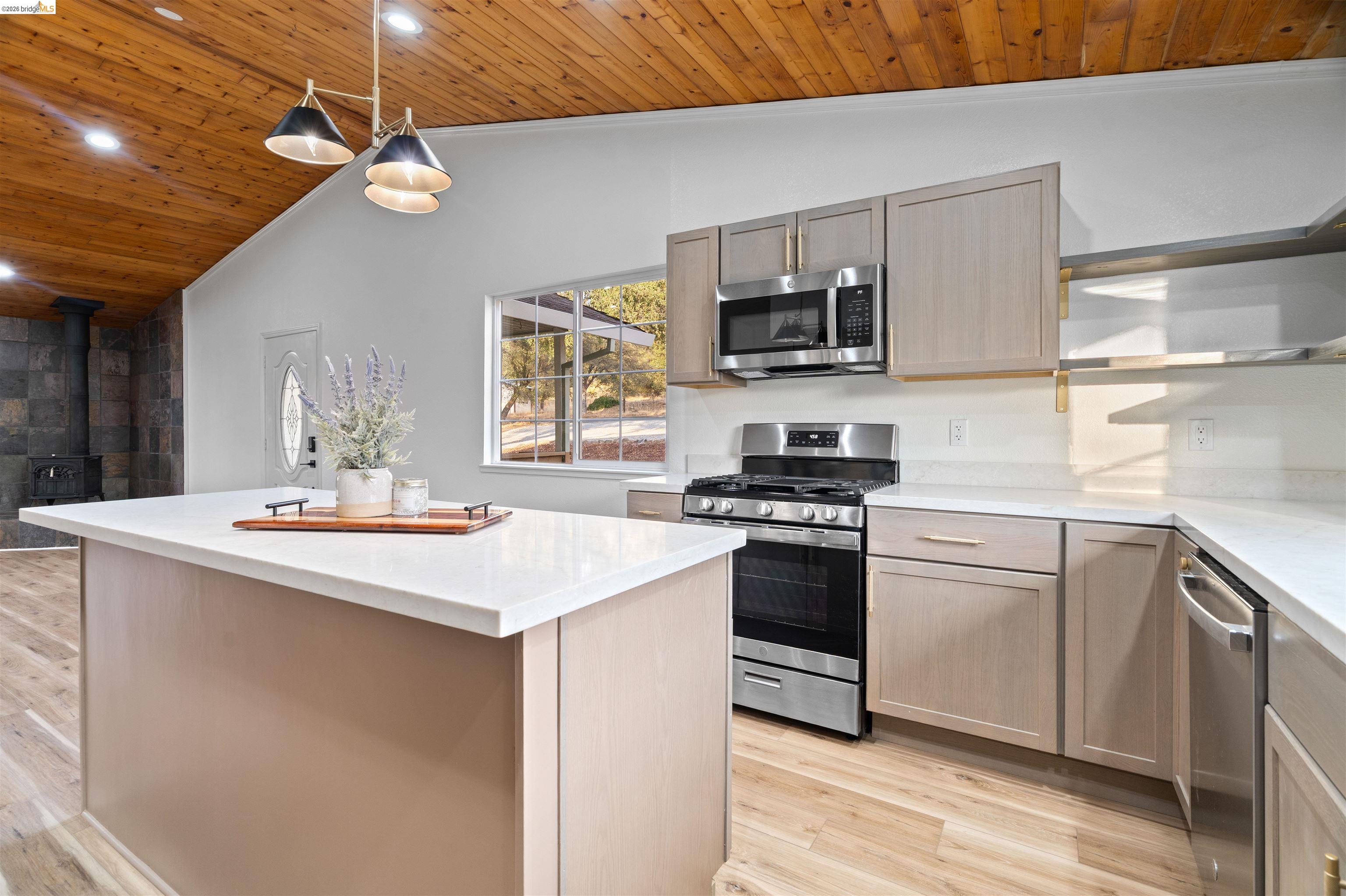 19676 Rawhide Road Sonora, CA 95370 - Photo 18 of 67 Contemporary kitchen featuring a large waterfall-edge island with white quartz countertops, light wood cabinetry, stainless steel appliances including a gas range and built-in microwave, pendant lighting, and wood-finish flooring throughout