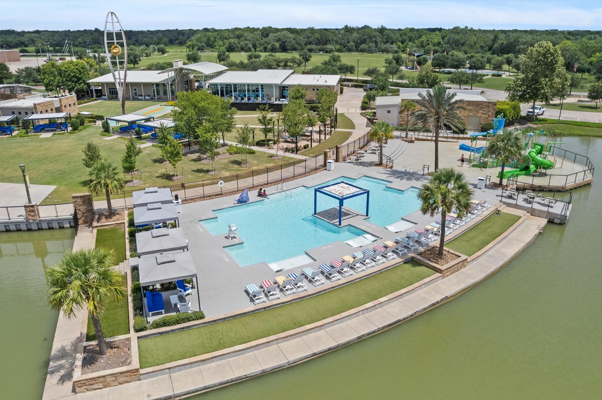 3510 Whitman Drive Rosharon, TX 77583 - Photo 20 of 25 This photo showcases a modern community pool area with ample lounge seating, shaded cabanas, and a waterslide. It's surrounded by lush greenery and offers a view of a nearby recreational building. Ideal for family-friendly living in a vibrant community setting.