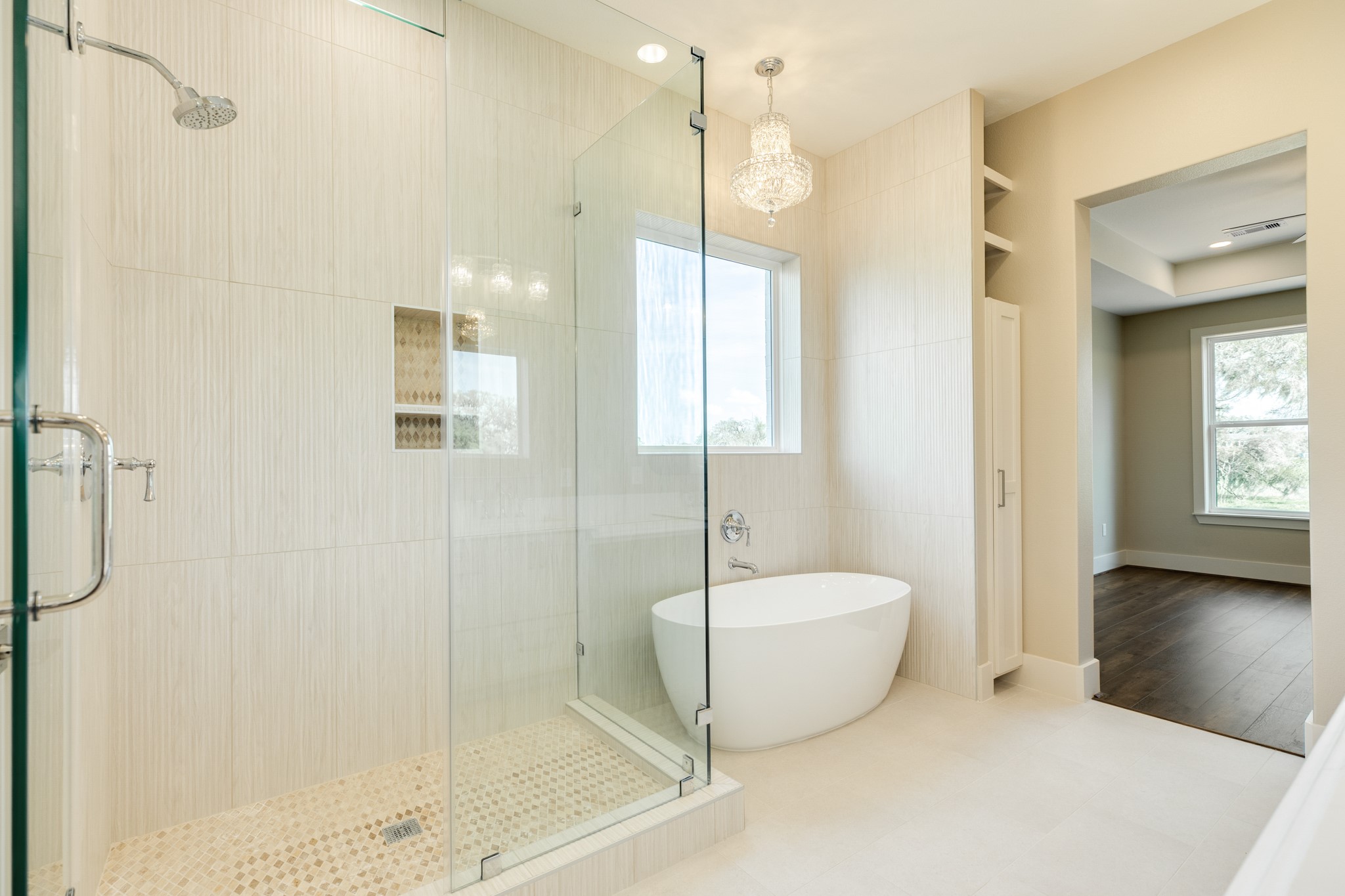 1737 Flintlock Court Angleton, TX 77515 - Photo 28 of 47 This photo showcases a modern bathroom featuring a spacious glass-enclosed shower and a freestanding bathtub. The room is well-lit with a large window and a chandelier, complemented by light-colored tiles and an adjacent area with hardwood flooring.
