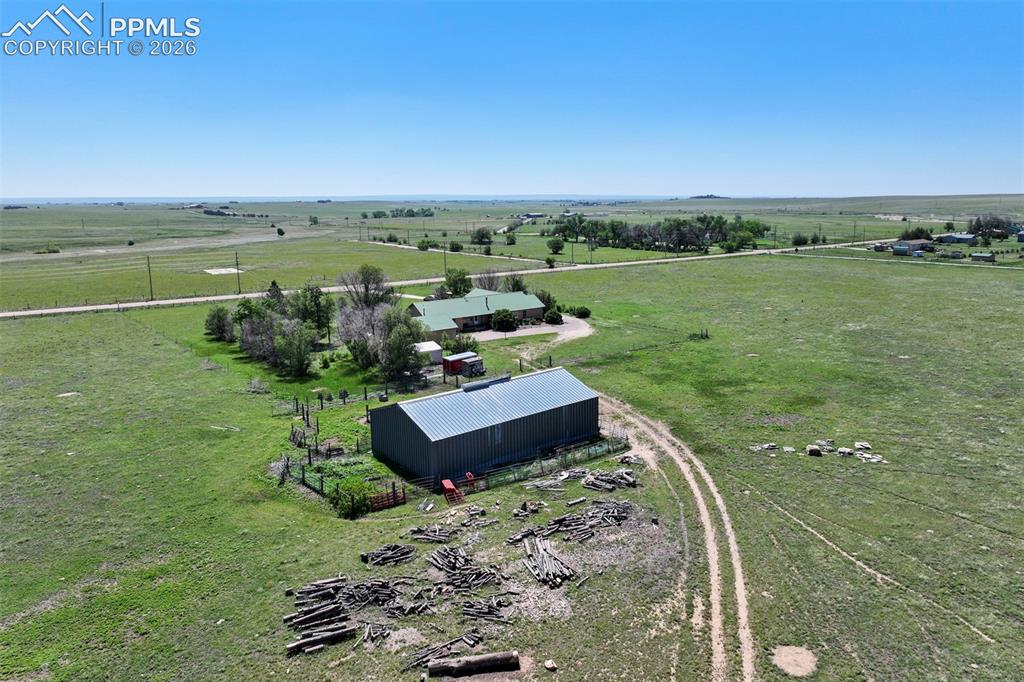 2550 Slocum Road Peyton, CO 80831 - Photo 5 of 50 Expansive rural property featuring a main residence with a green roof, a large metal outbuilding, a fenced garden area, mature trees, and a dirt road leading to the structures