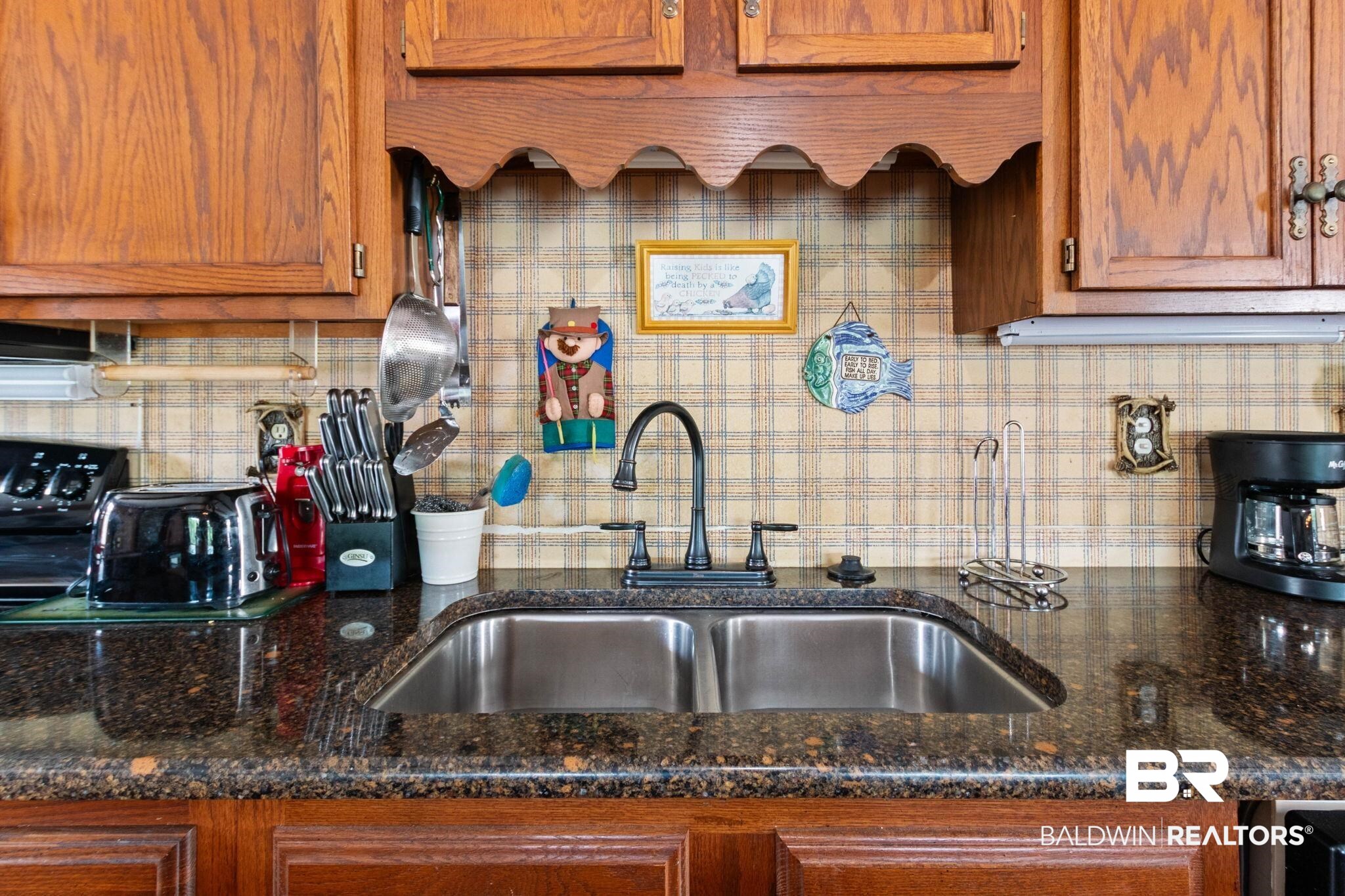 8153 Bryant's Lndg Road Stockton, AL 36579 - Photo 31 of 87 Kitchen featuring wood cabinetry, a double basin stainless steel sink, a dark polished countertop, a black pull-down faucet, and plaid wallpaper backsplash