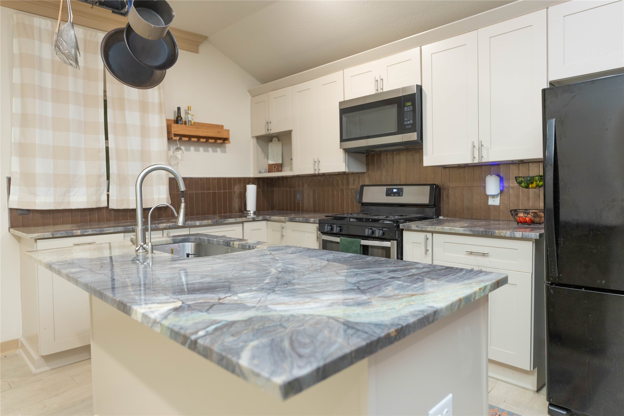 8007 Capitol Street Houston, TX 77012 - Photo 10 of 25 Modern kitchen with sleek marble countertops, white cabinetry, and stainless steel appliances. Features a central island with a sink, hanging pot rack, and contemporary tiled backsplash. Cozy and functional space for cooking and entertaining.