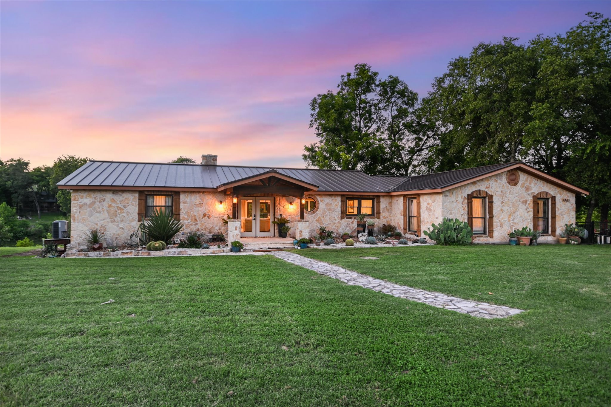 2411 Spoke Hollow Road Wimberley, TX 78676 - Photo 2 of 37 A stone-clad ranch house with a metal roof and an inviting entry, set against a vibrant sunset.