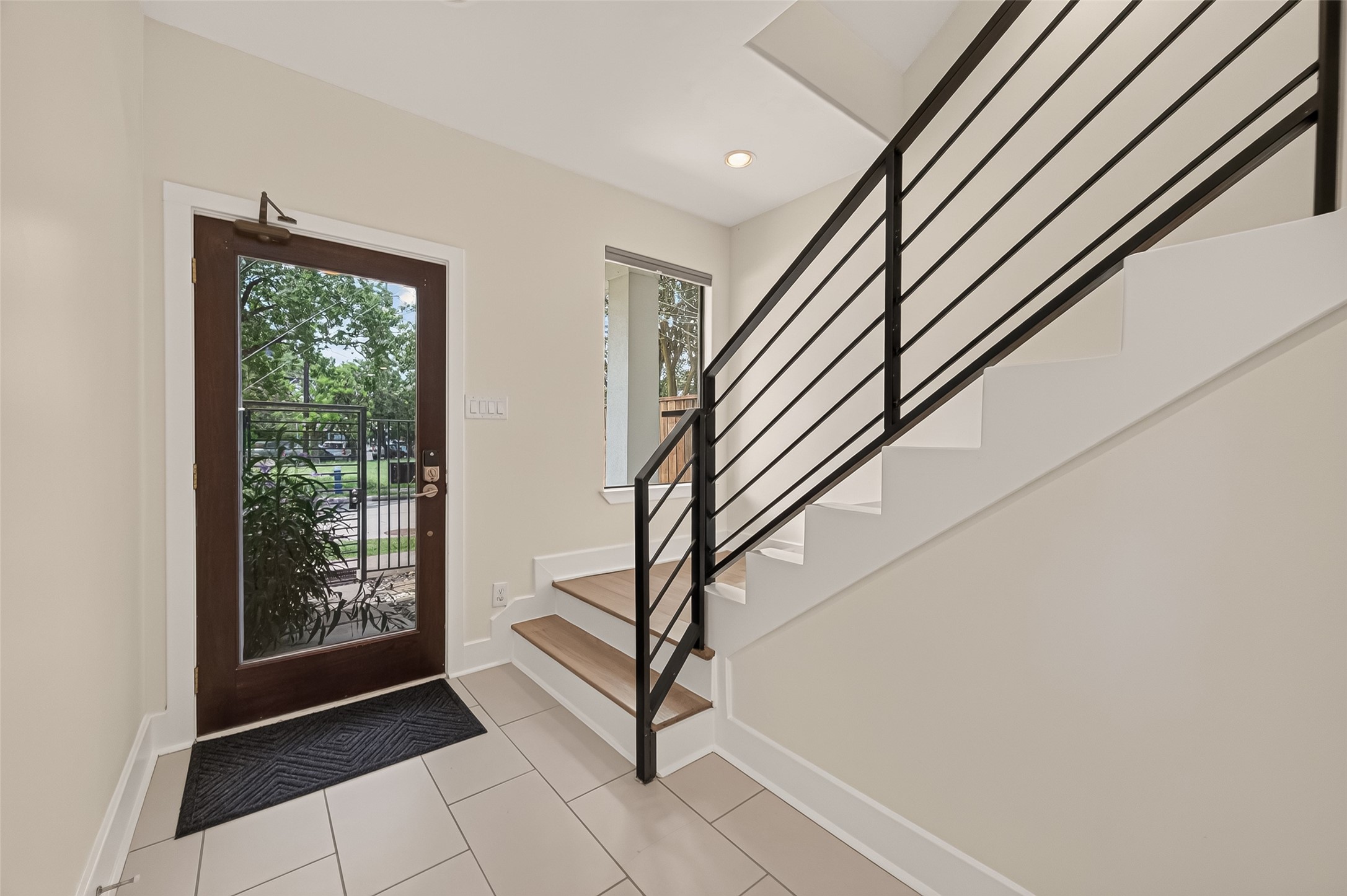 1767 Michigan Street Houston, TX 77006 - Photo 3 of 33 Bright entryway with a modern glass door, tile flooring, and a sleek staircase with a black metal railing. Large window offers natural light and a view of the greenery outside.