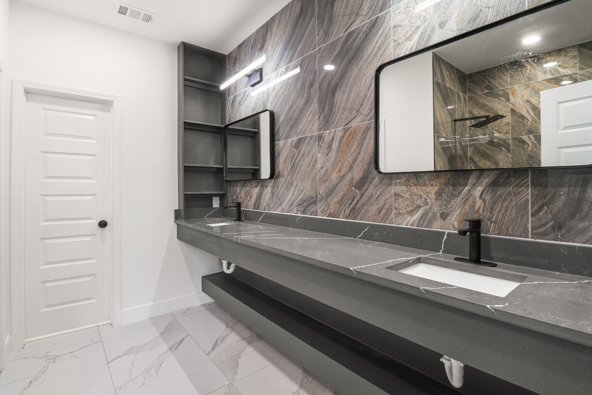 8046 Chateau Street Houston, TX 77028 - Photo 13 of 22 This bathroom features a modern double vanity with sleek black fixtures and a spacious dark countertop. The striking wall tiles add a touch of elegance, while the large mirrors enhance the room's brightness. Ample storage is available with open shelving, and the contemporary design is complemented by white and gray flooring.