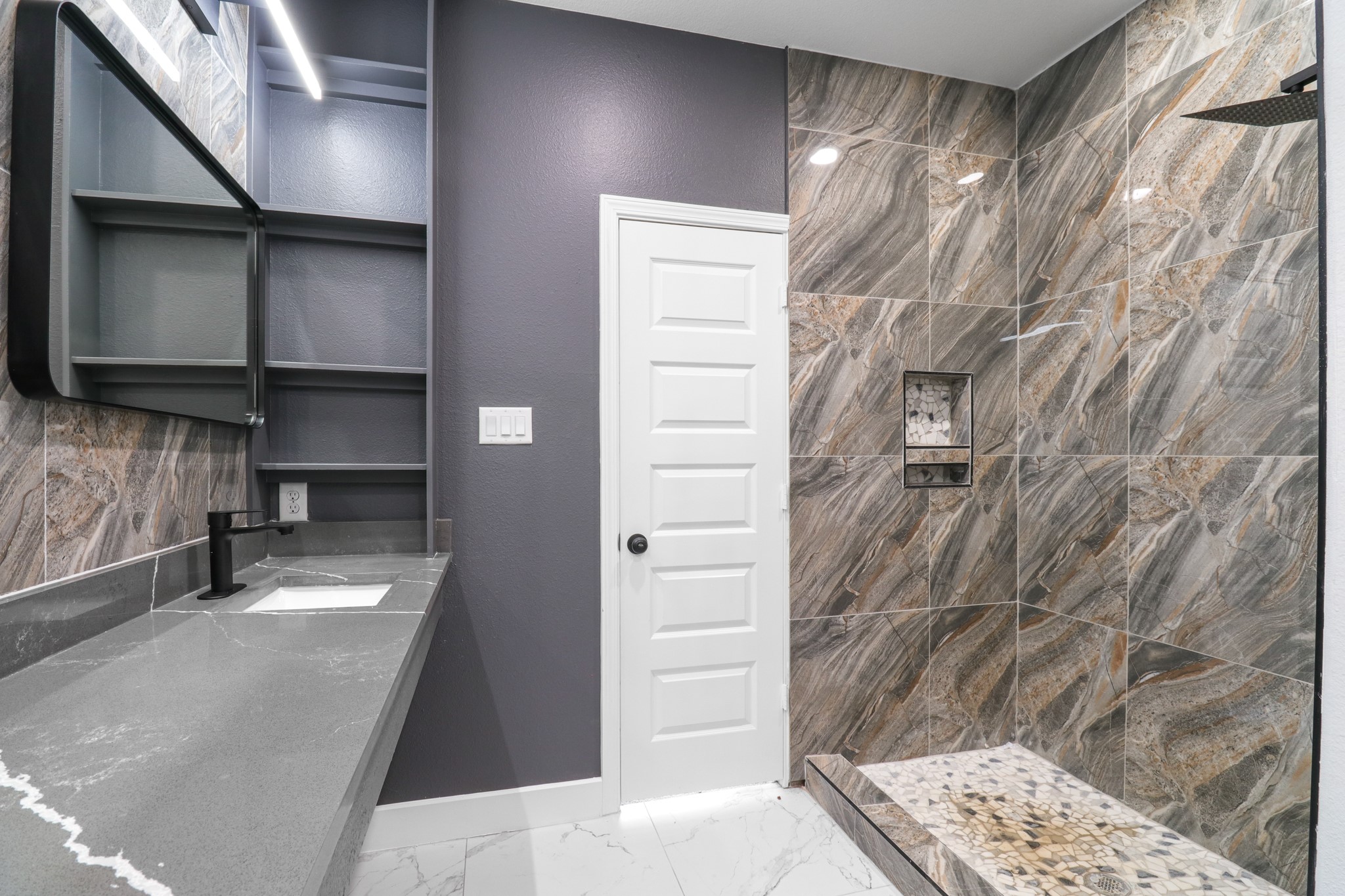 8046 Chateau Street Houston, TX 77028 - Photo 14 of 22 Modern bathroom with a sleek design featuring a large walk-in shower with textured stone tiles. It includes a spacious gray countertop with a built-in sink, open shelving, and stylish lighting. The room is accented with a neutral color palette, creating a sophisticated and inviting atmosphere.