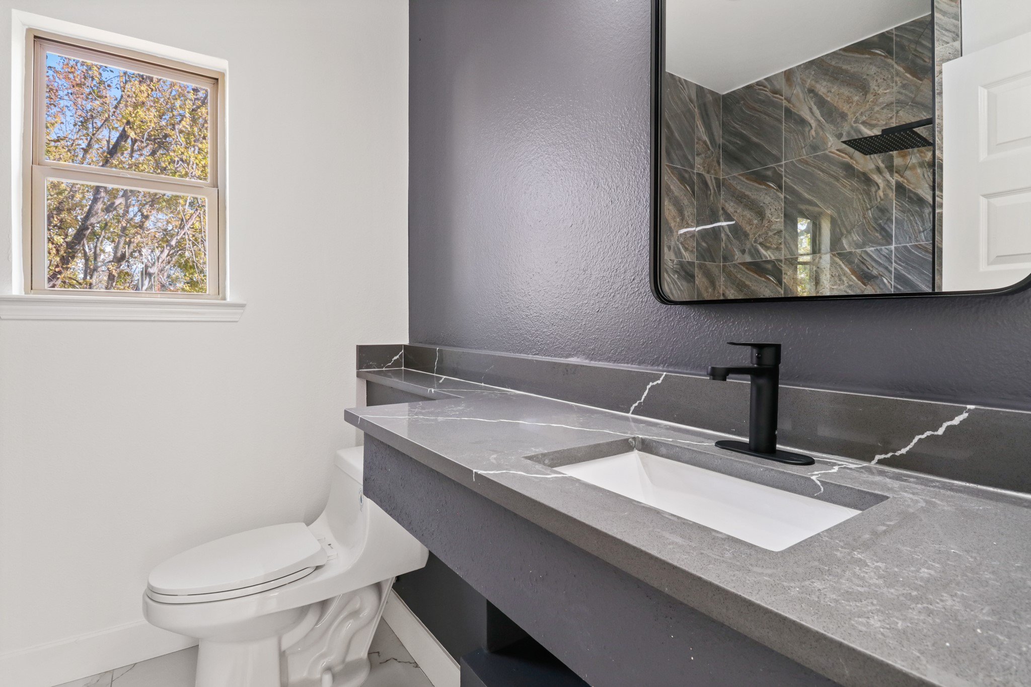 8046 Chateau Street Houston, TX 77028 - Photo 16 of 22 This modern bathroom features a sleek gray countertop with a stylish black faucet and a large mirror. The shower area is adorned with elegant stone tiles, and a window allows natural light to brighten the space.