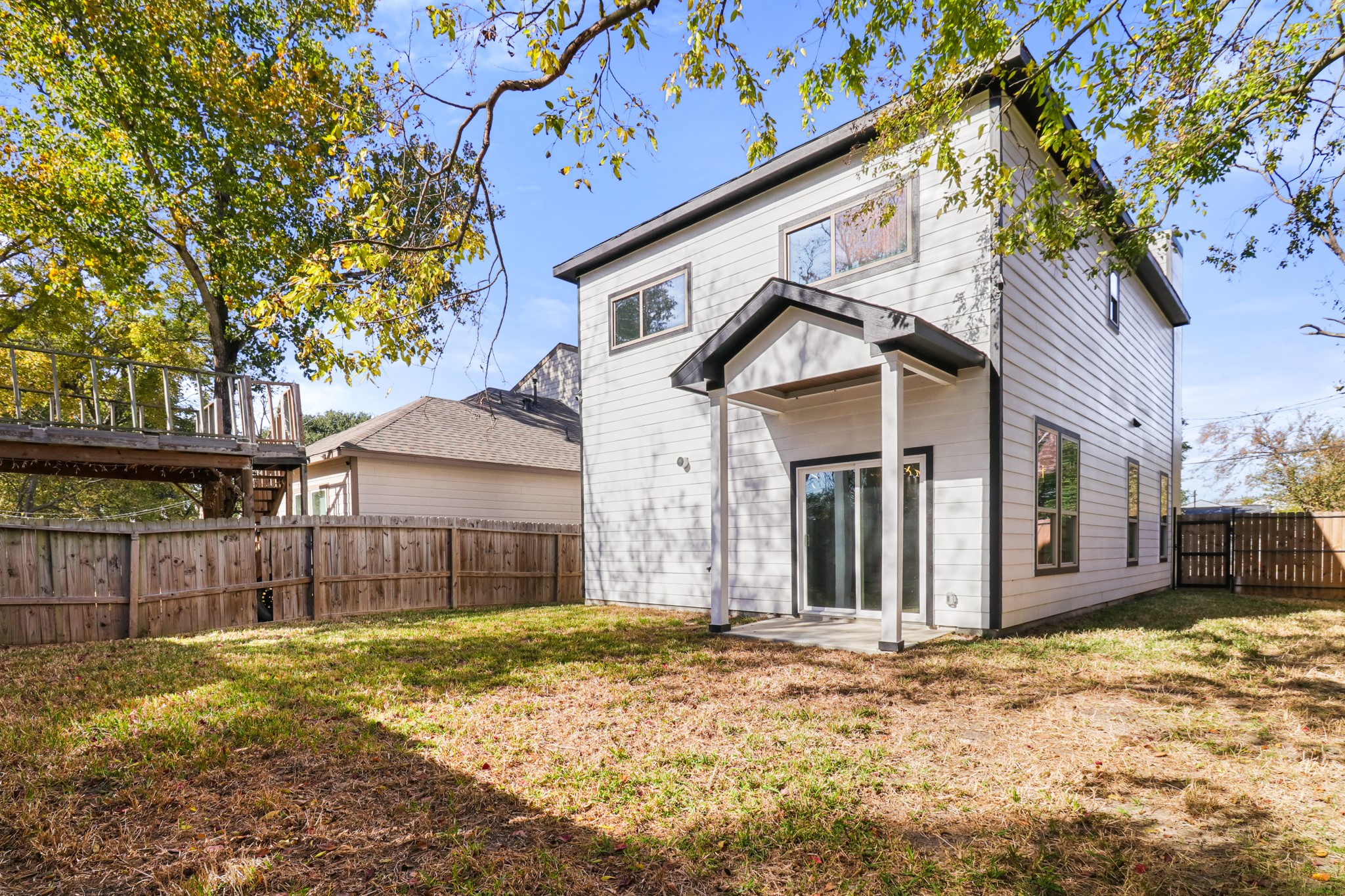 8046 Chateau Street Houston, TX 77028 - Photo 21 of 22 This photo shows a modern two-story house with a light exterior and a small covered patio. The backyard is fenced, providing privacy, and features both grass and mature trees, offering a blend of sun and shade. Ideal for outdoor activities or relaxation.