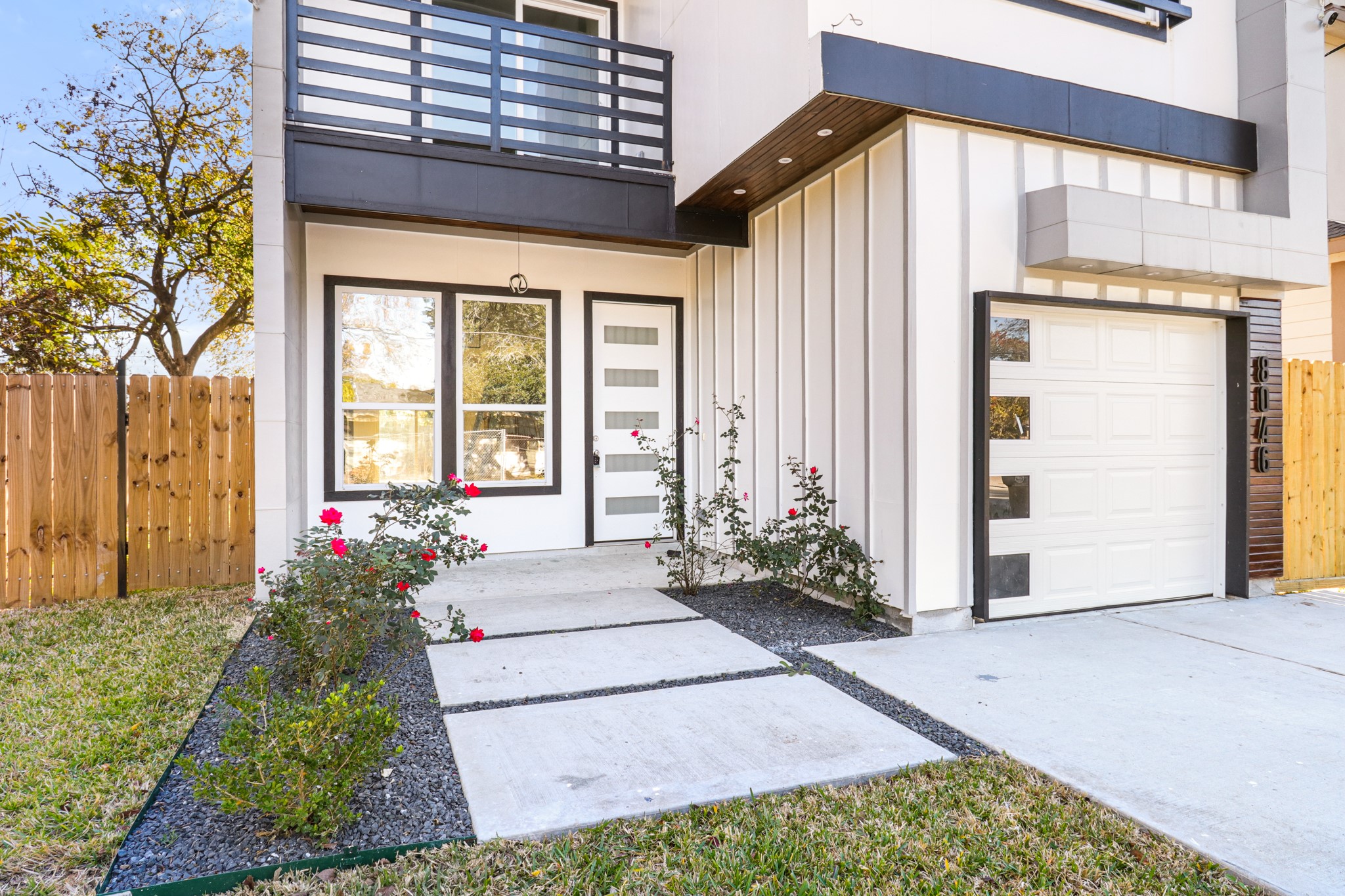 8046 Chateau Street Houston, TX 77028 - Photo 4 of 22 Modern two-story home with a sleek facade, featuring large windows and a balcony. The entrance is framed by a small garden with vibrant flowers, and there's a single-car garage. A wooden fence encloses the side yard, providing privacy.