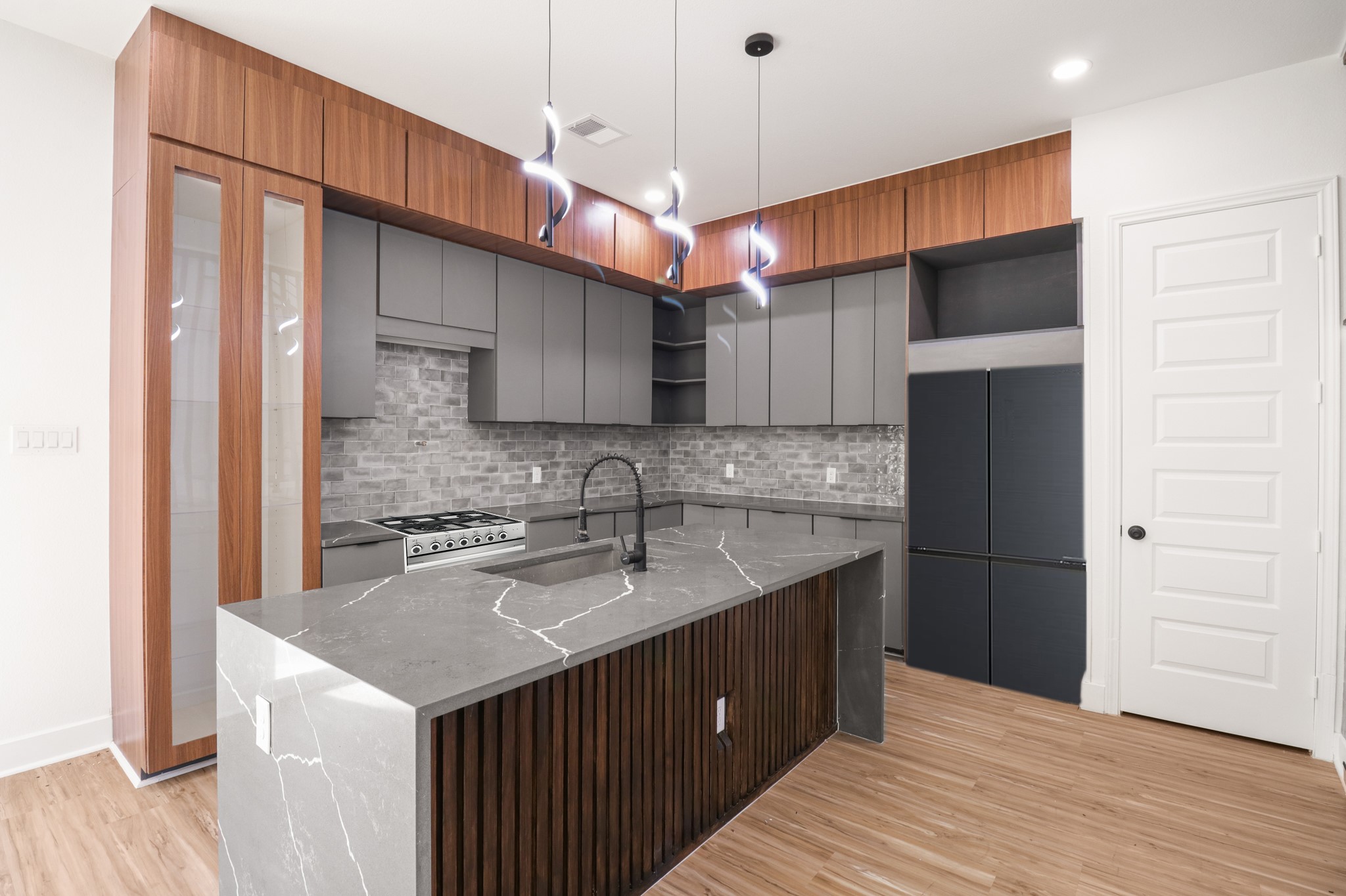 8046 Chateau Street Houston, TX 77028 - Photo 6 of 22 Modern kitchen featuring sleek gray cabinetry, elegant wood accents, and a stylish marble countertop island. The room is illuminated by contemporary pendant lights, with a brick-style backsplash adding texture. Perfect for a sophisticated, functional space.