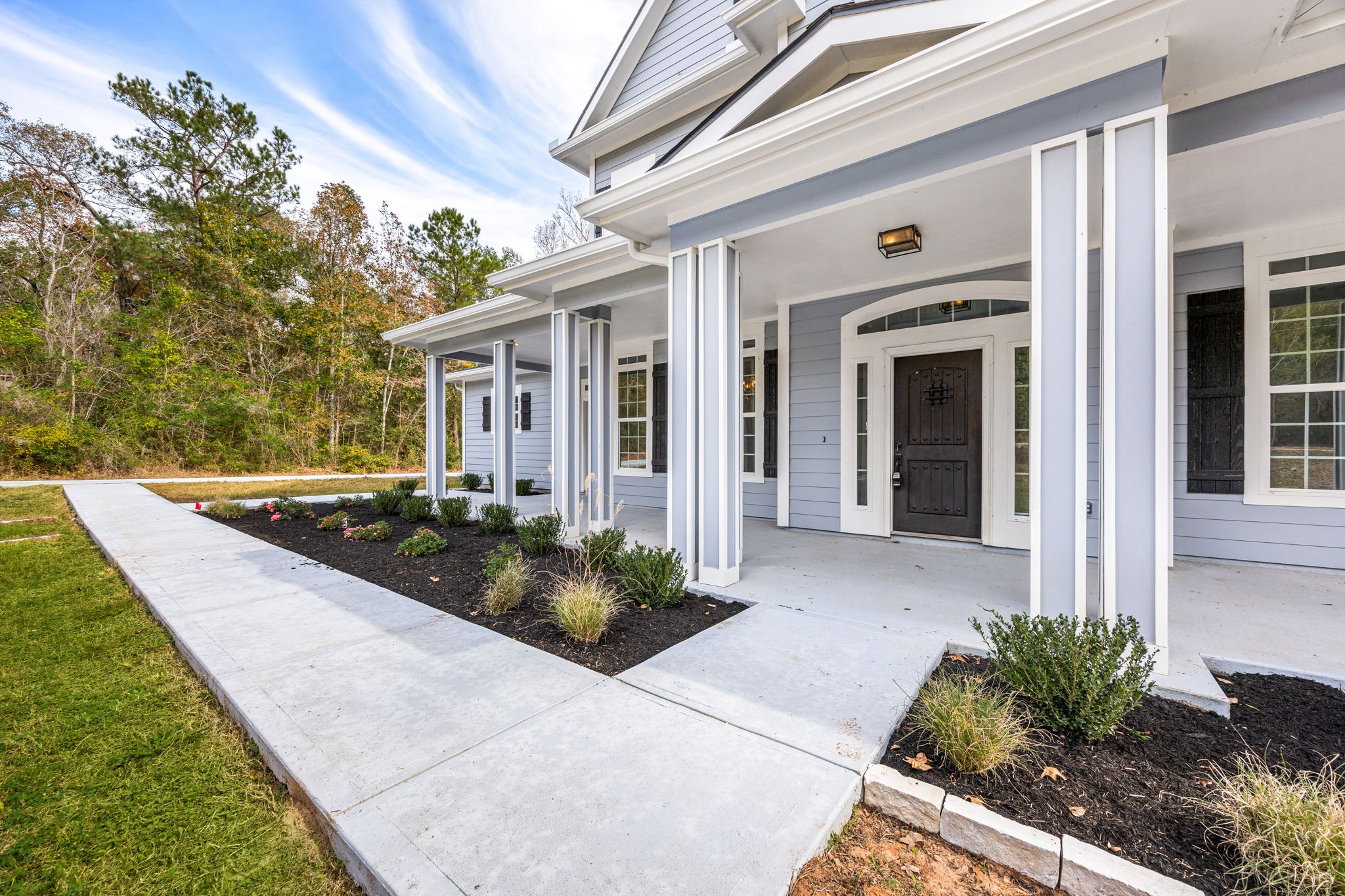 14882 Gingham Road Montgomery, TX 77356 - Photo 2 of 41 A welcoming porch, featuring clean lines and a neatly landscaped front yard. The house is painted in a soft blue-gray with white trim, providing a fresh and inviting appearance. The surrounding area includes a paved walkway and lush greenery, adding to the home's curb appeal.