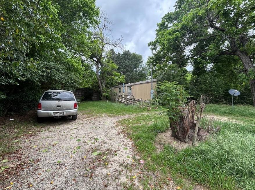 Property featuring a gravel driveway, mature trees, and a tan-sided structure with an exterior utility meter