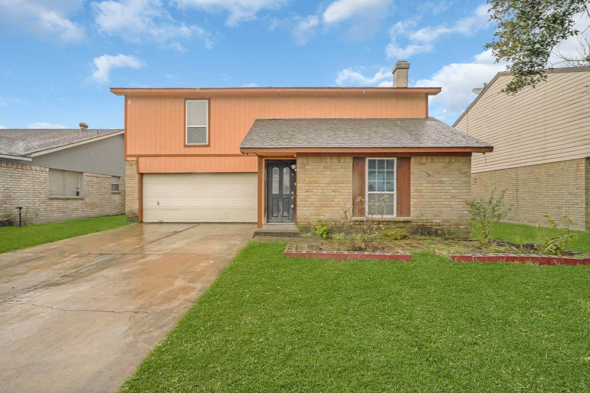 This is a two-story home with a brick and wood exterior. It features a two-car garage and a well-maintained front lawn with a small garden bed. The driveway is spacious, and the house has a welcoming entrance.
