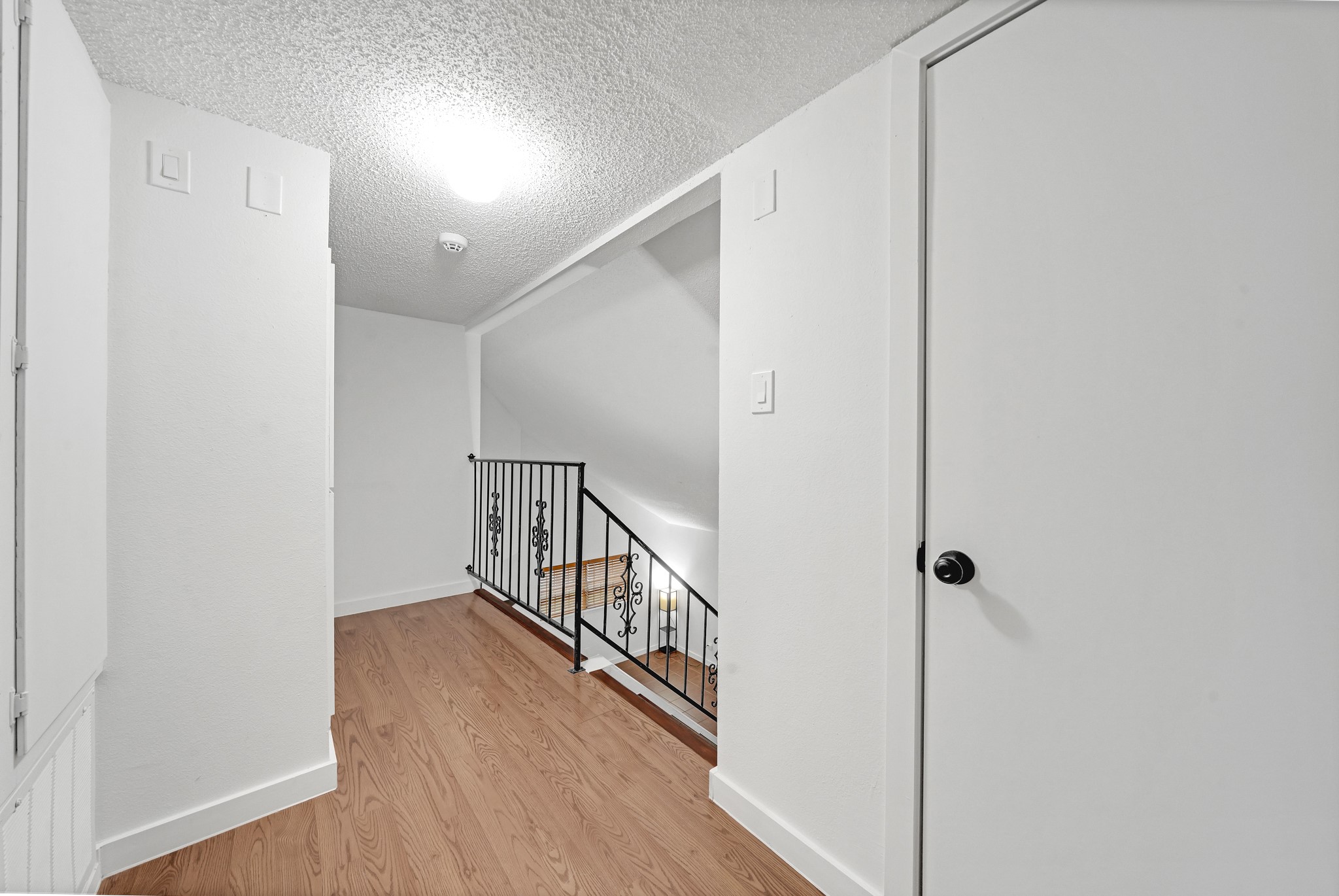 635 Walston Lane Houston, TX 77060 - Photo 14 of 24 This photo shows a bright hallway with wood flooring, white walls, and a textured ceiling. There's a staircase with a decorative black metal railing leading down, and a closed door to the right, suggesting access to another room or storage.
