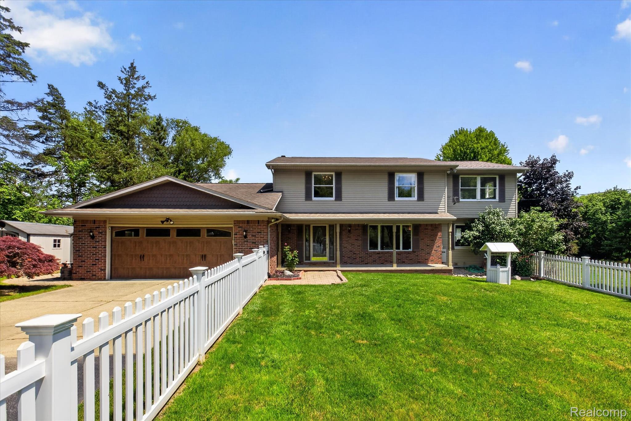 7186 East River Road Flushing, MI 48433 - Photo 1 of 47 Two-story residence featuring a brick and siding exterior with dark brown shutters, an attached two-car garage with a wood-finish door, a manicured front lawn, and a white picket fence