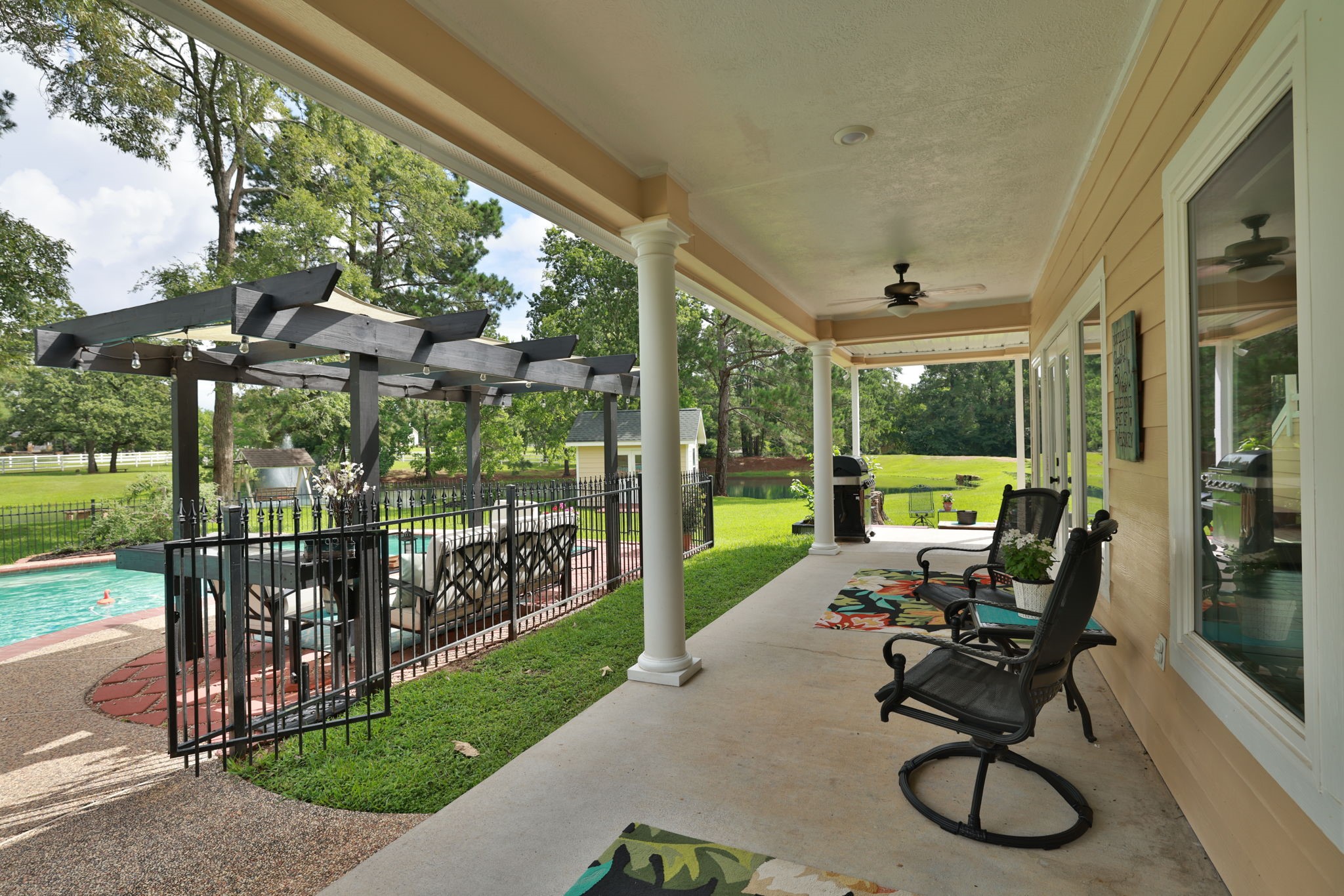 34310 High Point Drive Magnolia, TX 77355 - Photo 36 of 48 This photo showcases a spacious covered patio with ceiling fans, overlooking a fenced pool area with a pergola and seating. It offers a serene view of a lush green yard, perfect for outdoor relaxation and entertaining.