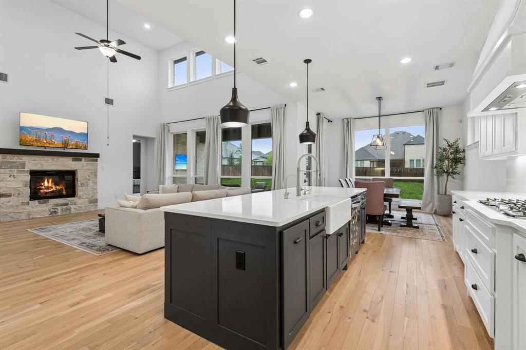 566 Broadleaf Way Haslet, TX 76052 - Photo 3 of 38 Open-concept living space featuring a spacious kitchen island with a farmhouse sink, light wood-finish flooring, a stone-surround fireplace with a dark wood mantle, high vaulted ceilings, and a dining area with outdoor access