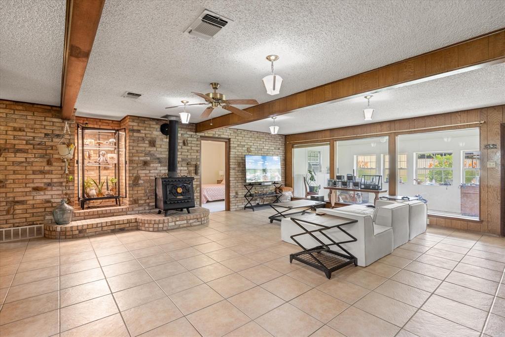 4210 Waples Road Granbury, TX 76049 - Photo 17 of 37 Spacious living area featuring light-toned tile flooring, exposed wood beams, and a brick accent wall with an integrated wood-burning stove on a raised hearth