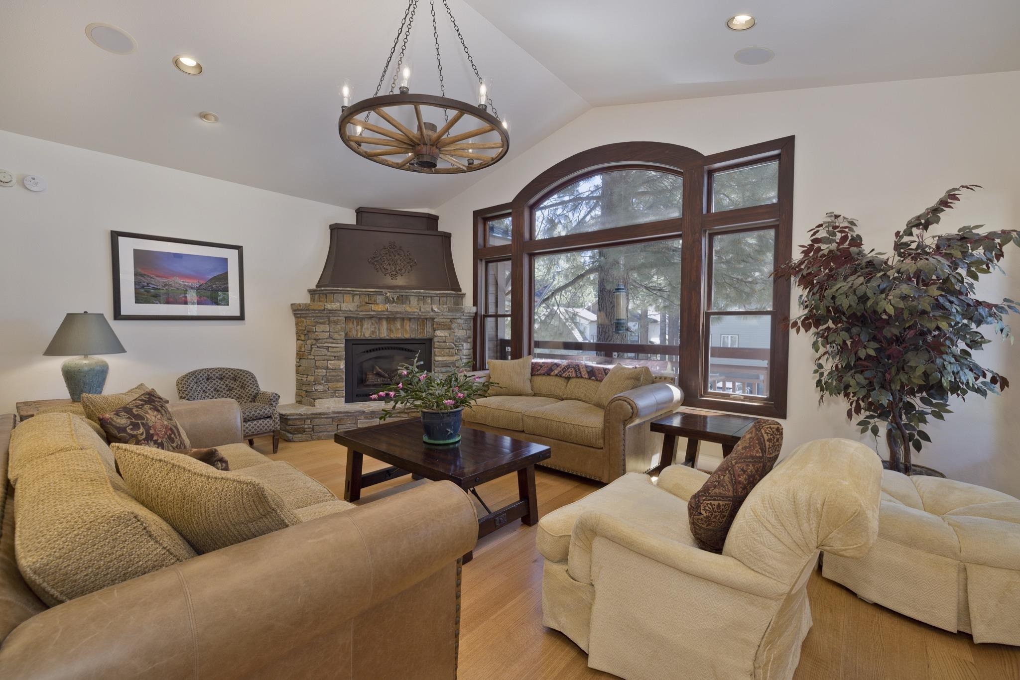 378 Ridge Way Mammoth Lakes, CA 93546 - Photo 7 of 43 Vaulted ceiling living area featuring a stone-clad fireplace with a custom metal hood, large arched window, wood-finish flooring, and a wagon wheel chandelier