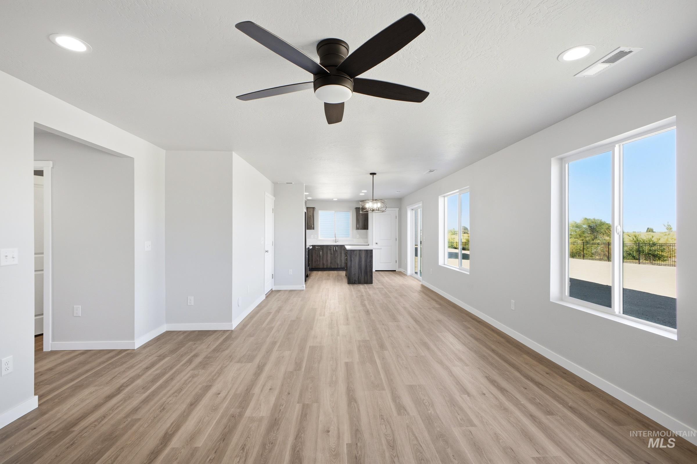 13325 Scrape Street Caldwell, ID 83607 - Photo 9 of 24 Spacious open-concept living area showcasing wood-finish flooring throughout, abundant natural light from expansive windows, contemporary ceiling fan with integrated lighting, and seamless sightlines to the kitchen and dining spaces beyond