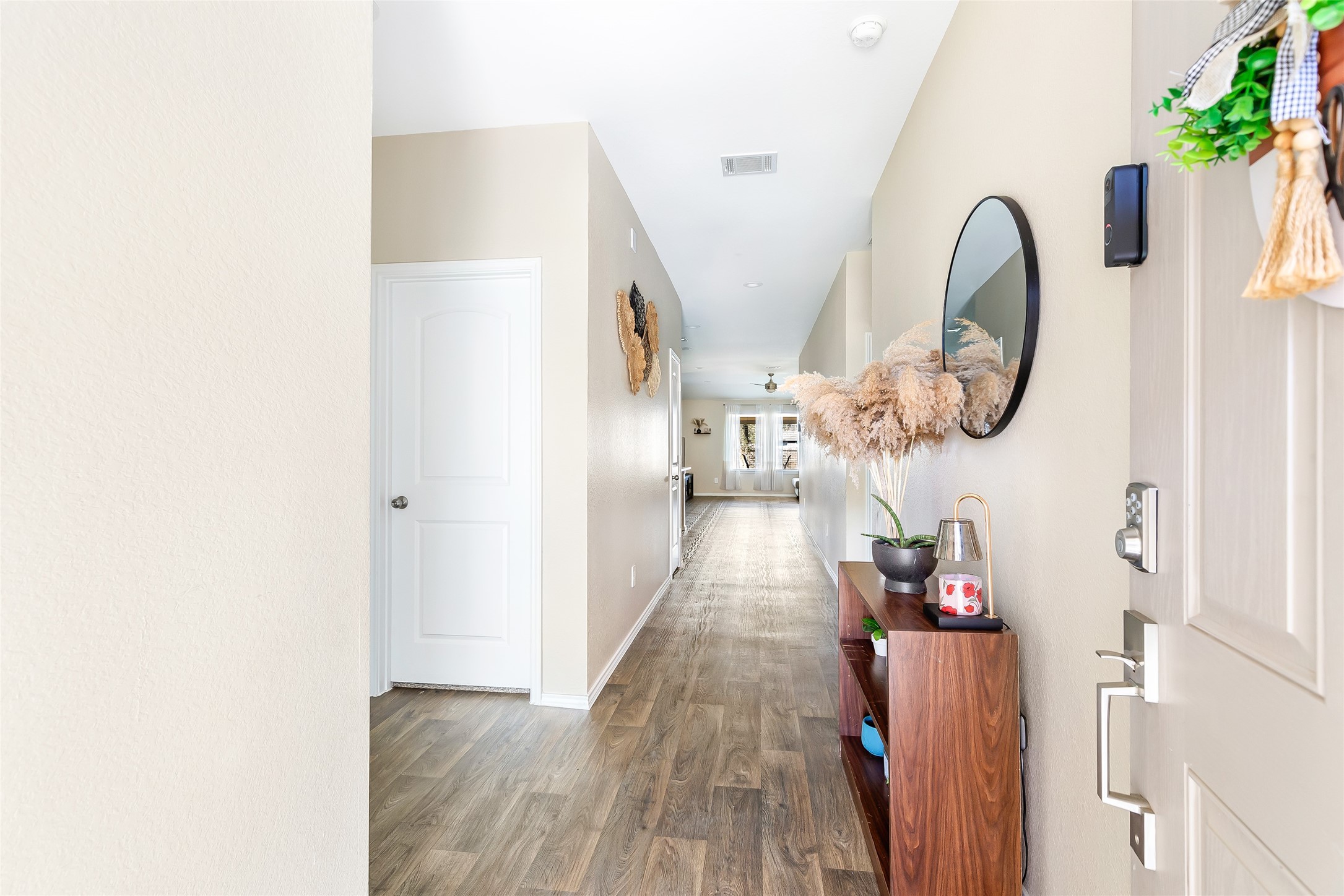 811 Republic Road New Caney, TX 77357 - Photo 14 of 37 This photo showcases a bright, welcoming hallway with light-color walls and wood like flooring. It features a small console table with decor and a round mirror, leading towards an open, airy living space with natural light at the end.