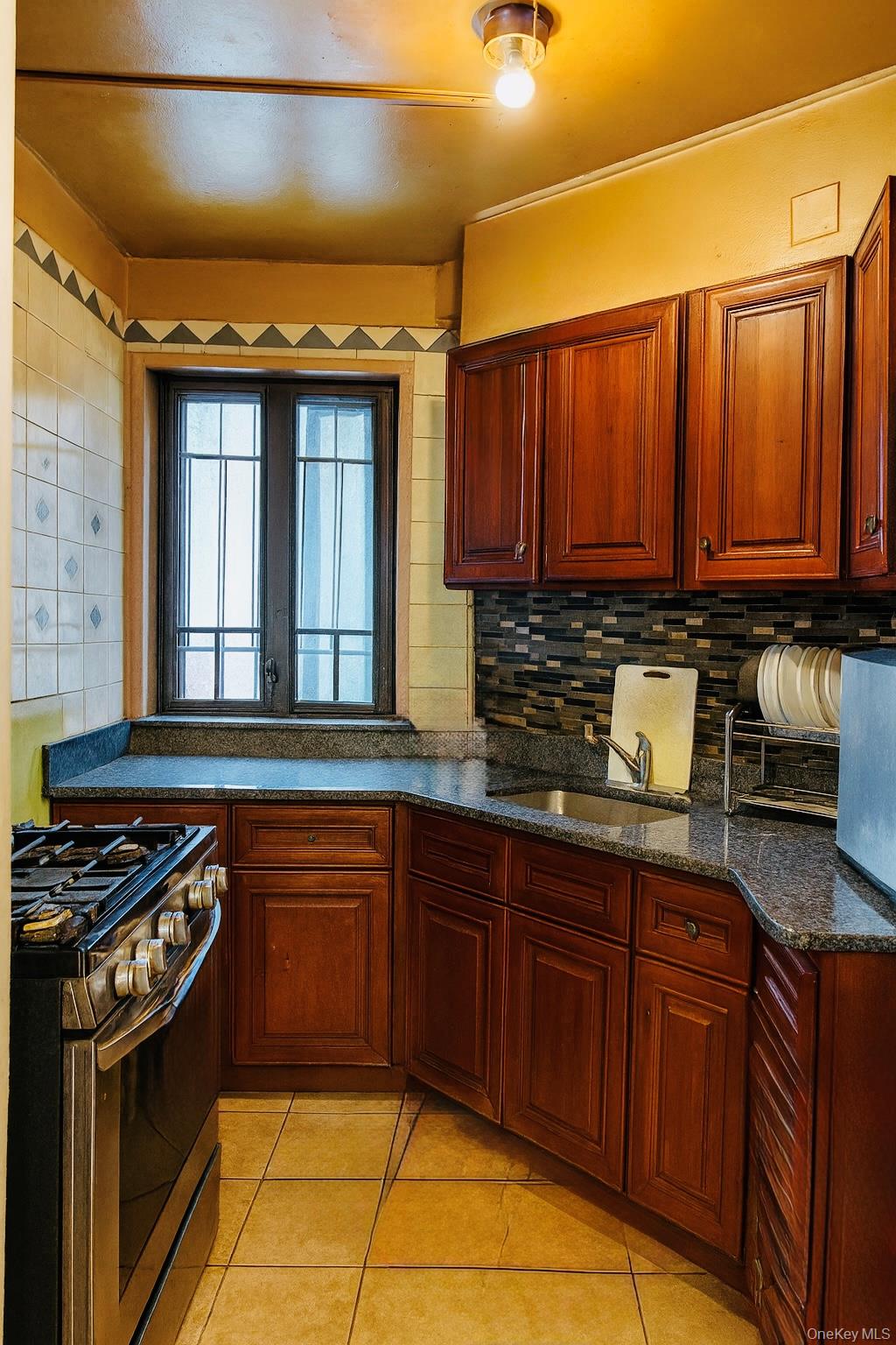 Kitchen featuring stainless steel gas range oven, light tile patterned floors, dark stone countertops, backsplash, and reddish brown cabinets