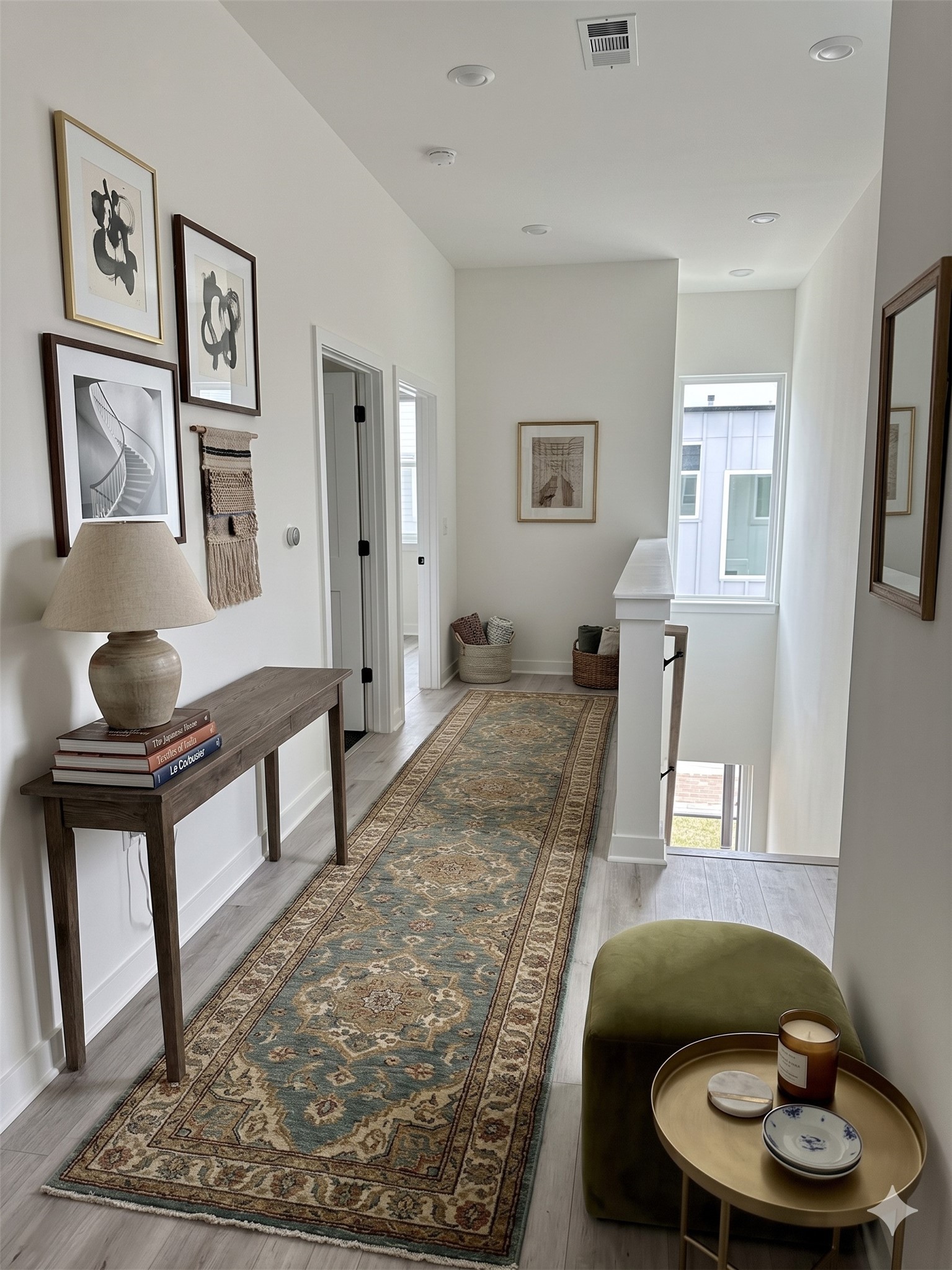 2000 Riverside Farms Road, Unit 19102 Austin, TX 78741 - Photo 4 of 13 This photo showcases a bright, stylish hallway with light wood flooring and white walls. It features a long, patterned rug and a wooden console table adorned with a lamp and books. There are framed artworks on the walls and a green ottoman with a side table holding candles. The space is well-lit with natural light from a window at the end.