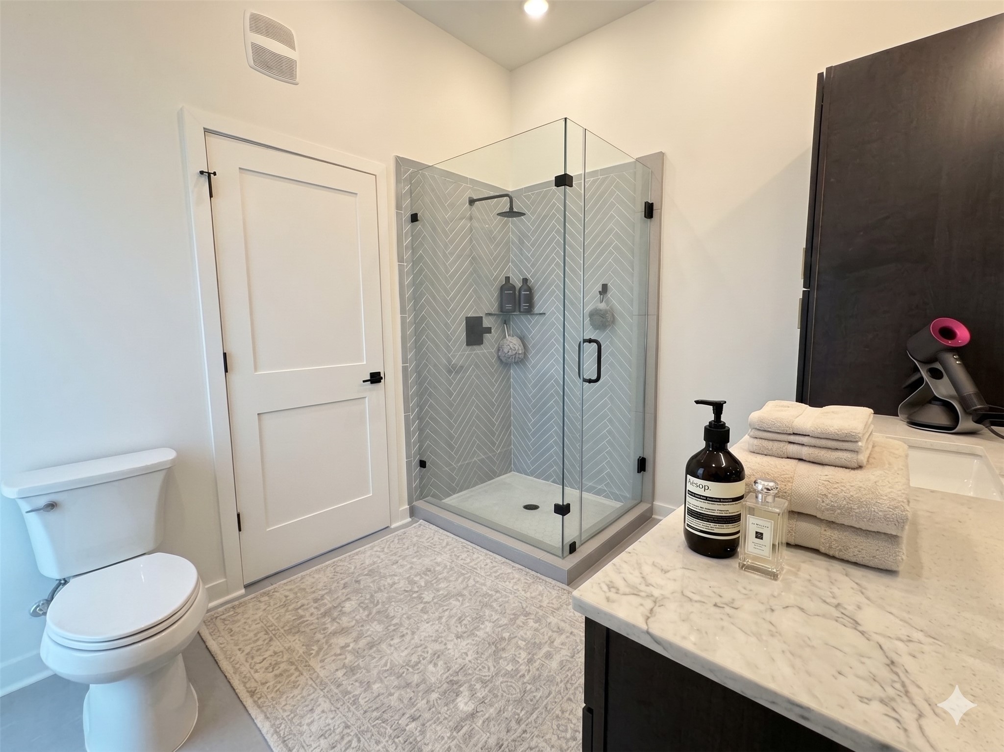 2000 Riverside Farms Road, Unit 19102 Austin, TX 78741 - Photo 7 of 13 This modern bathroom features a glass-enclosed shower with elegant chevron-patterned tiles, a sleek vanity with marble countertop, and a neatly arranged countertop with towels and toiletries, creating a clean and inviting space.