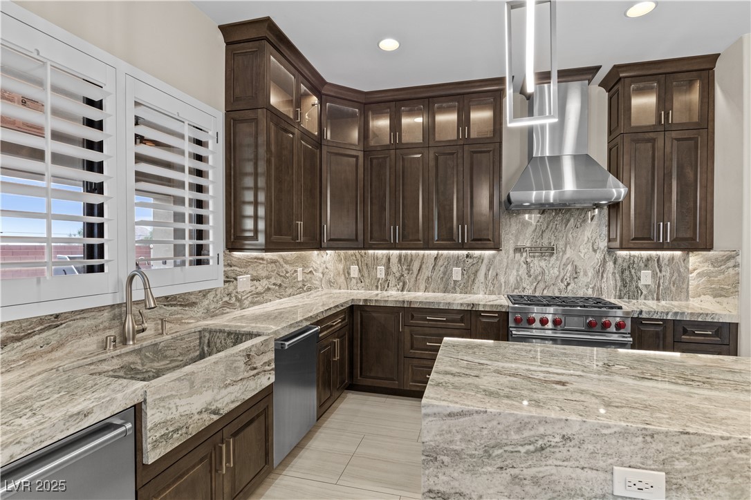 9865 Bright Angel Way Las Vegas, NV 89149 - Photo 24 of 98 Kitchen featuring wall chimney range hood, stainless steel appliances, decorative backsplash, dark brown cabinets, and light stone countertops